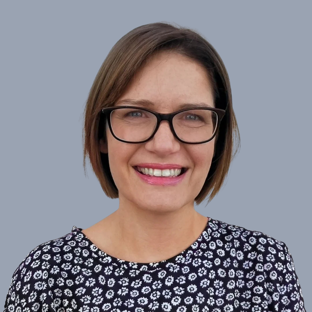 A woman with short brown hair, wearing glasses, smiling, and dressed in a black and white floral blouse against a light blue background.
