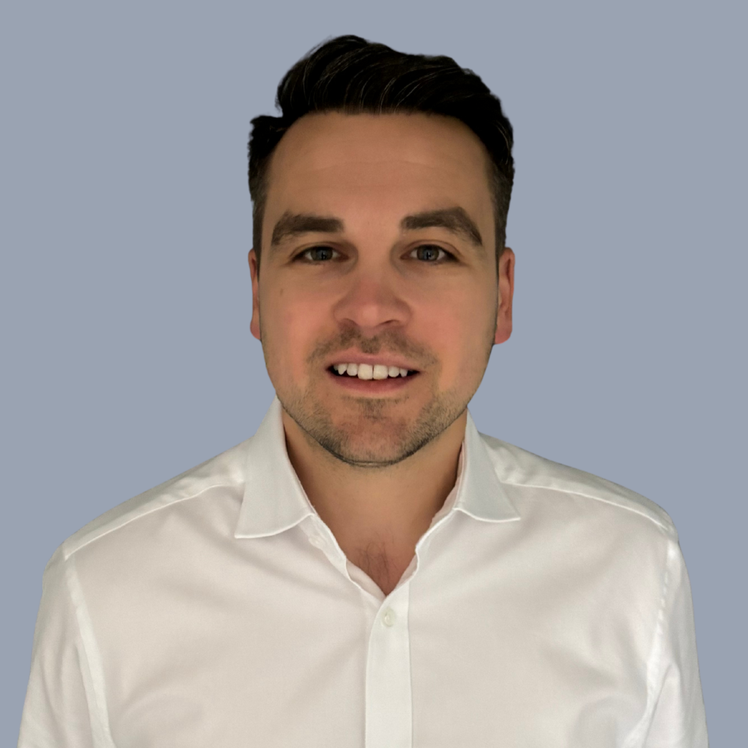 A young man with dark hair, wearing a white shirt, smiling at the camera against a light gray background.