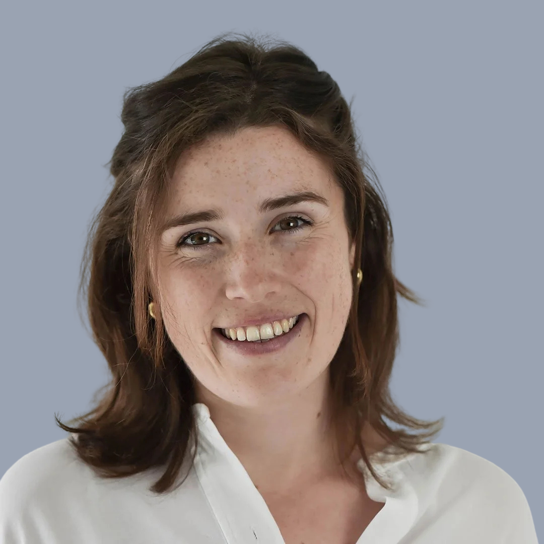 A woman with shoulder-length brown hair, wearing a white top and gold earrings, smiling against a plain gray background.