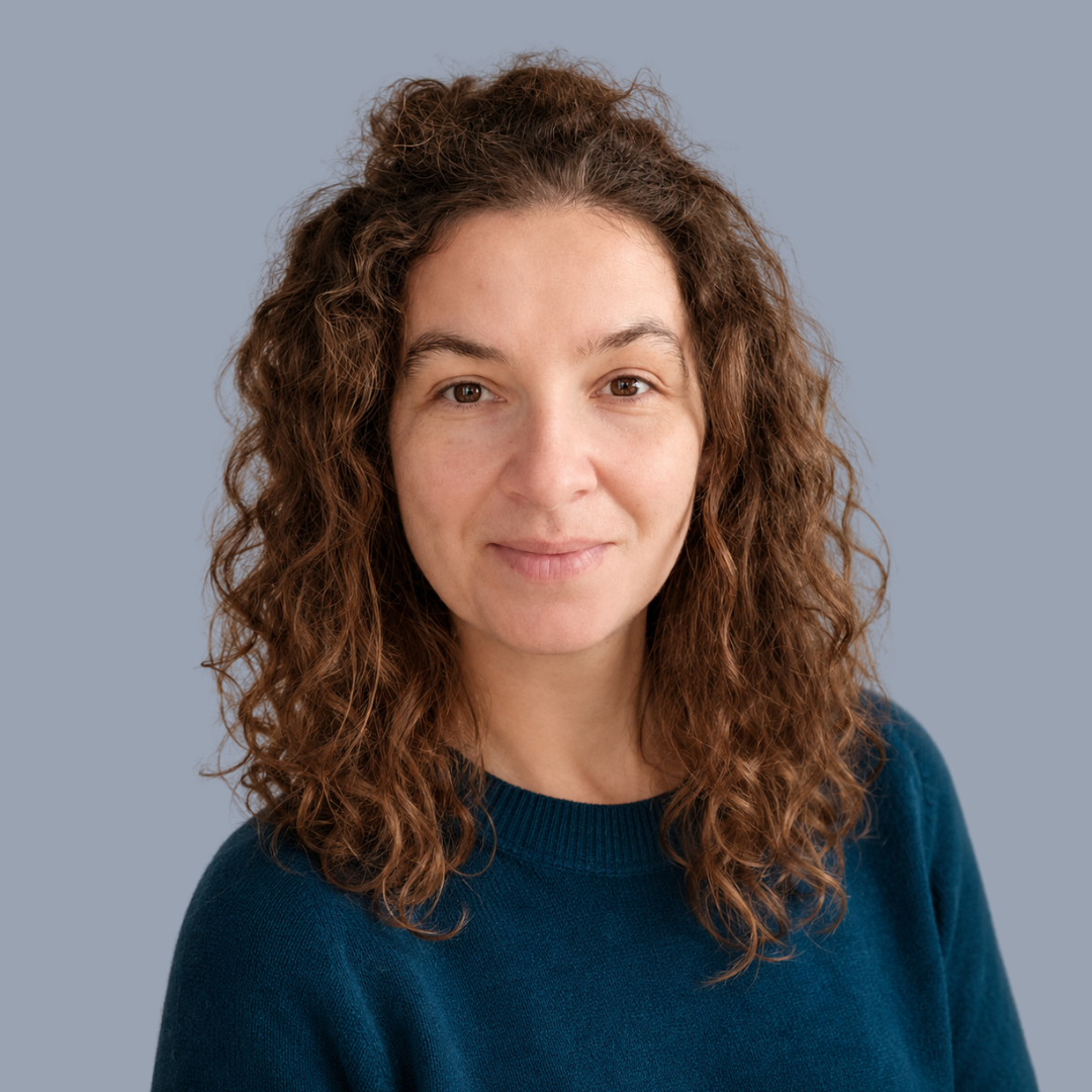 Portrait of a woman with curly brown hair wearing a navy blue sweater against a light grey background.