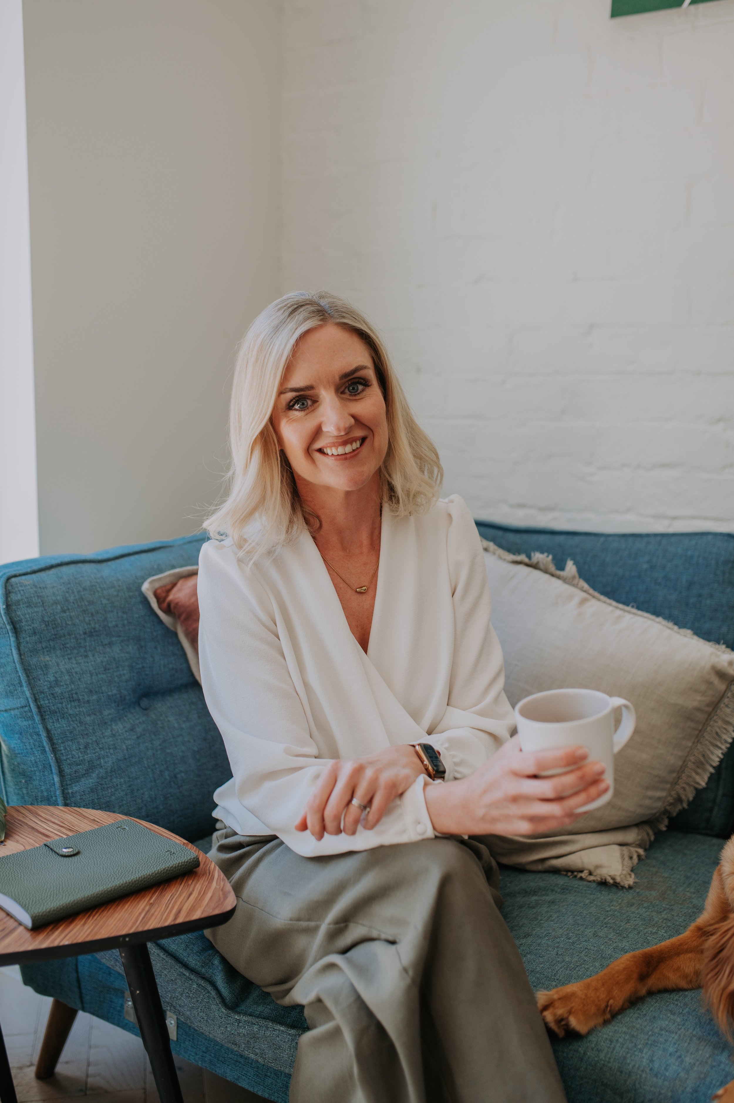 A woman with blonde hair smiling, sitting on a blue couch holding a white mug, with a beige pillow behind her. She is wearing a white blouse, khaki pants, a watch, and a necklace. There is a wooden table with a green notebook and a suitcase next to her, in a room with white brick walls.
