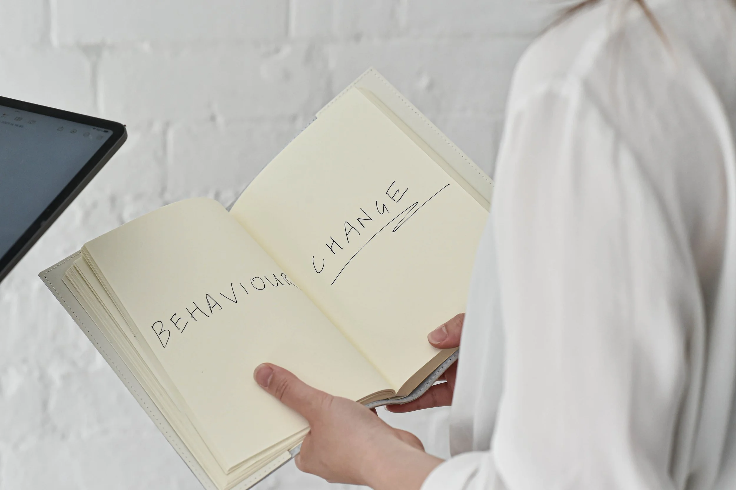 Person holding an open notebook with the words "BEHAVIOUR CHANGE" written on the pages.