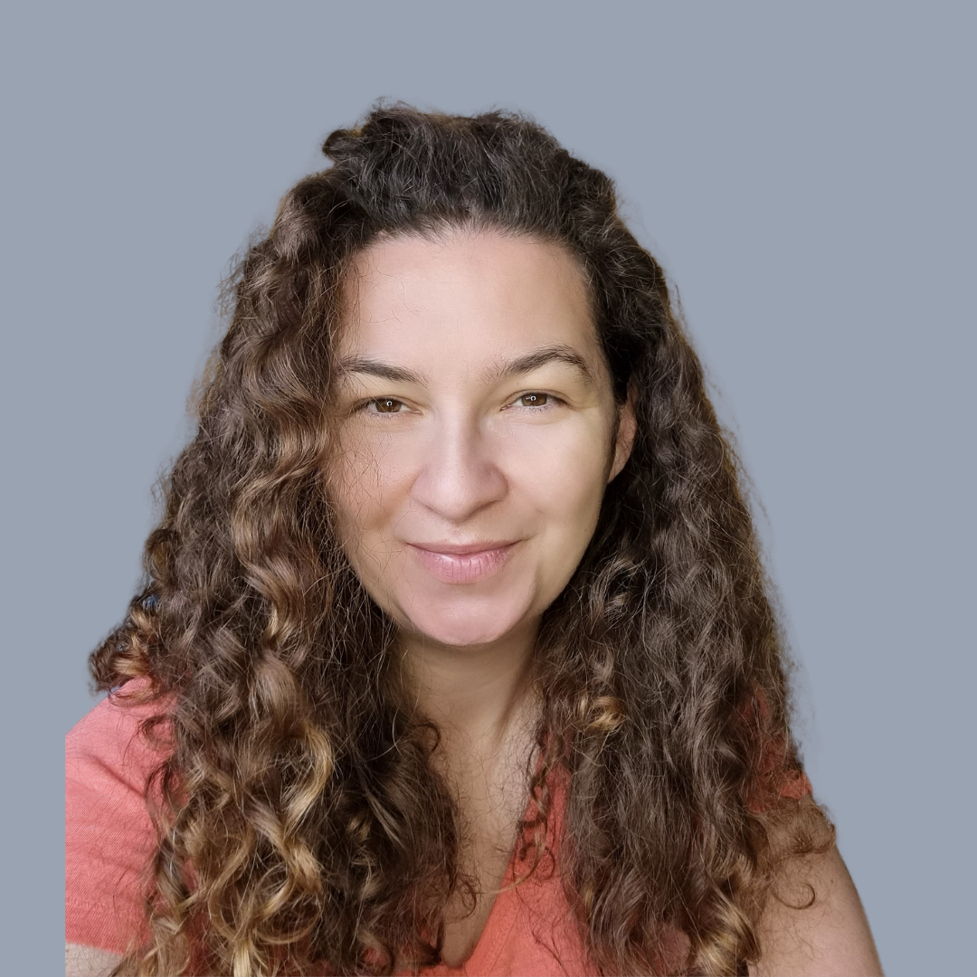 A woman with long, curly brown hair and light skin, smiling softly, wearing a coral top, against a gray background.