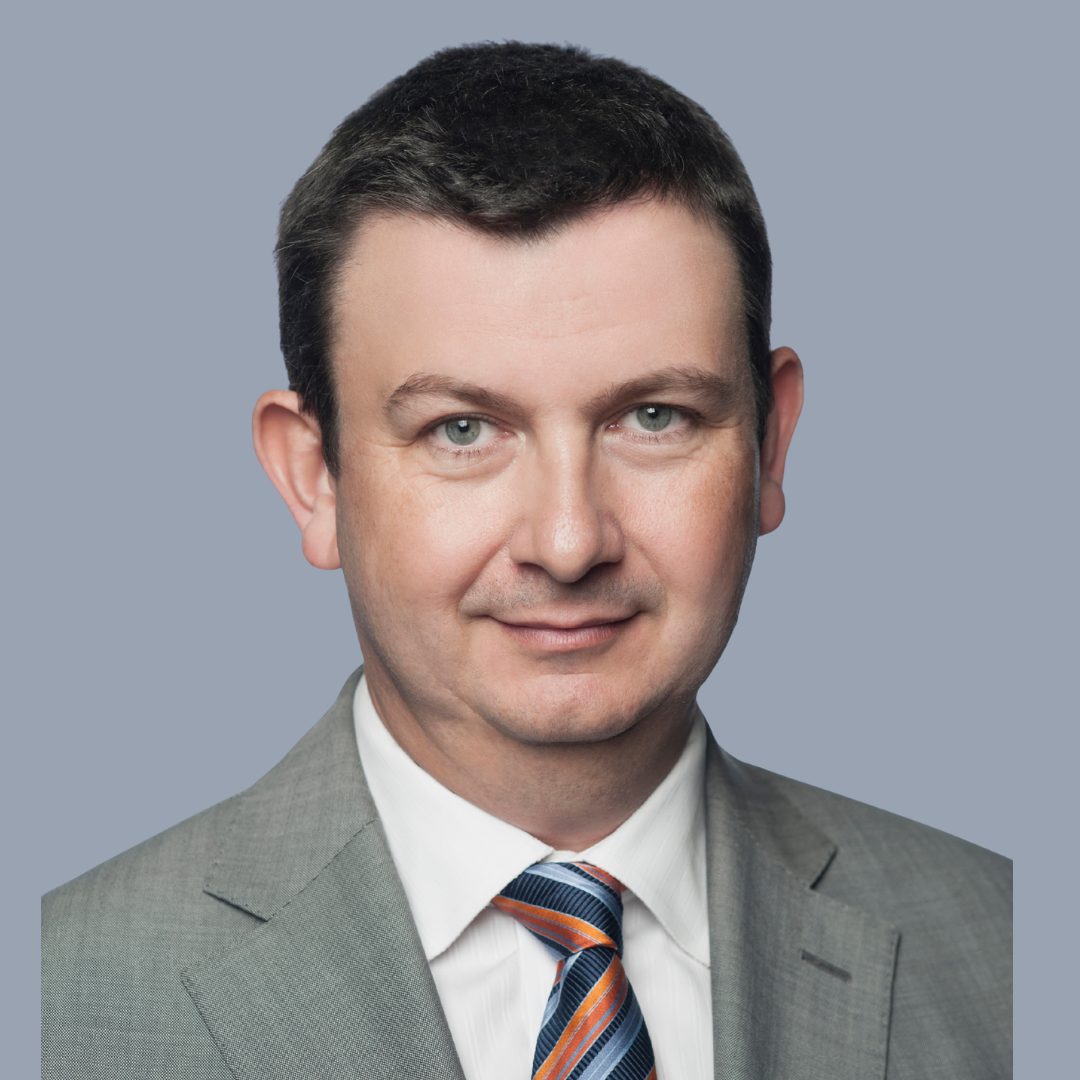 Professional headshot of a man in a gray suit, white shirt, and striped tie, smiling with a neutral gray background.