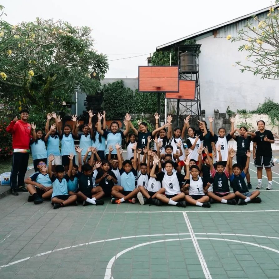 Group of children and their coach on an outdoor basketball court, some children in light blue shirts and others in black and white jerseys, posing for a team photo with their hands raised.