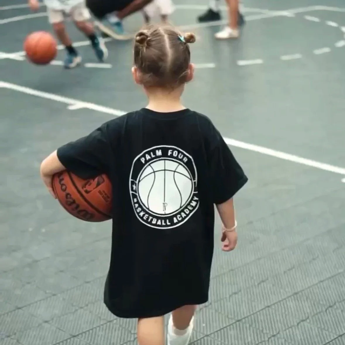 A young girl with braided hair holding a basketball in a gym. She is wearing a black T-shirt with the Palm Four Basketball Academy logo on the back, and is heading towards a basketball game in the background.