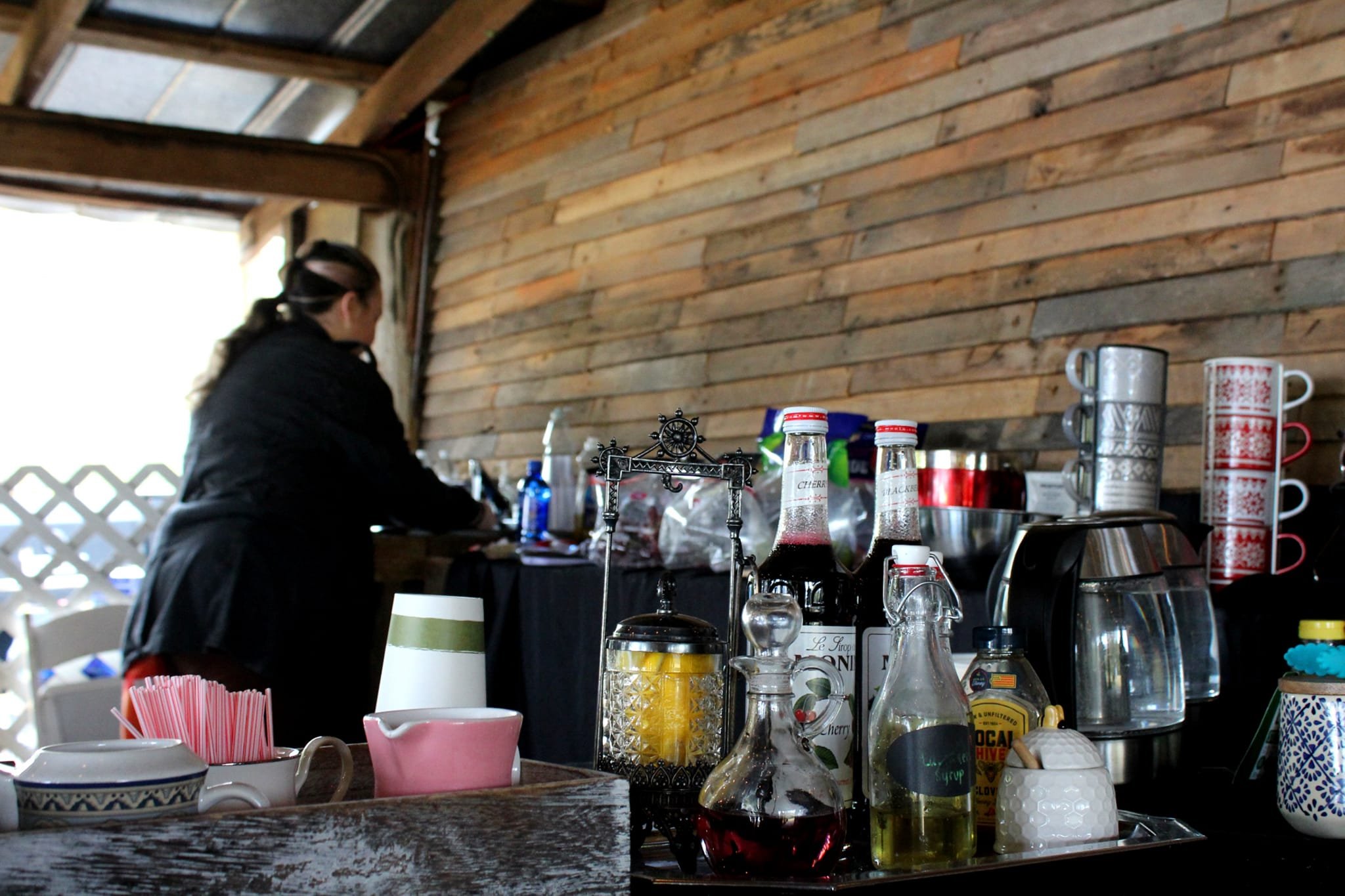 Back view of a woman in black jacket working behind a bar with various bottles and mugs on a wooden counter