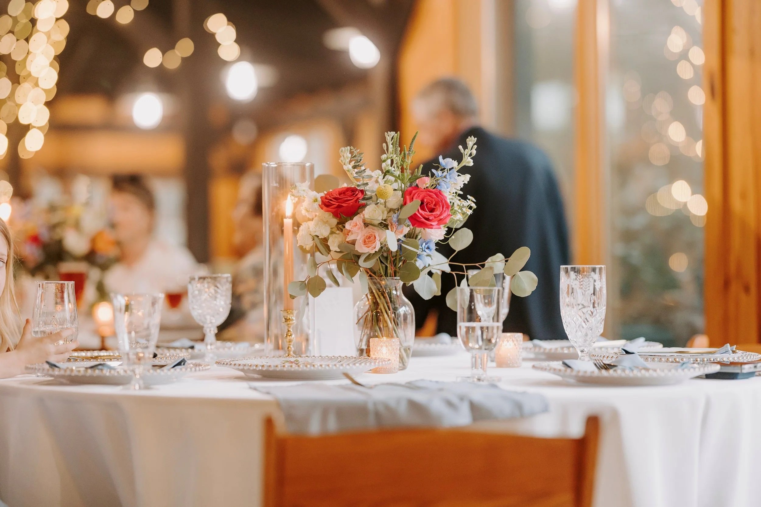 A wedding reception table decorated with a floral centerpiece, candles, and elegant glassware, with blurred guests and warm lighting in the background.
