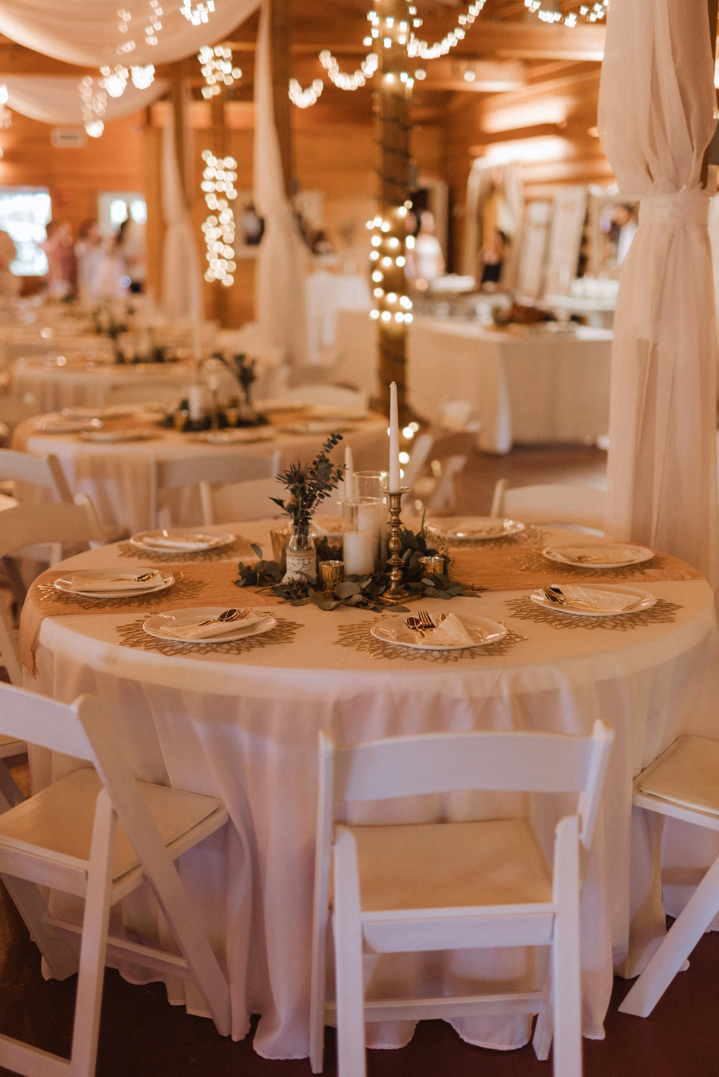 A decorated banquet hall with round tables covered in white tablecloths, set with plates, silverware, and napkins. Each table features a centerpiece with candles, greenery, and gold accents. The room has string lights and draped fabric overhead, creating a warm, festive atmosphere.