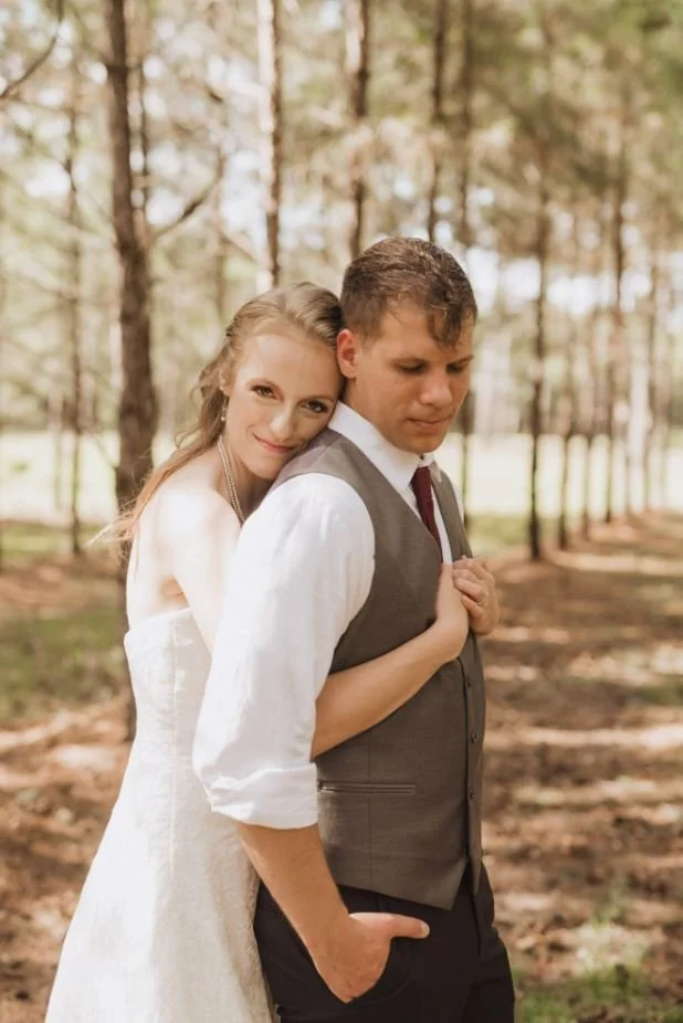 A woman in a wedding dress hugging a man in a vest and dress shirt, standing outdoors in a wooded area.