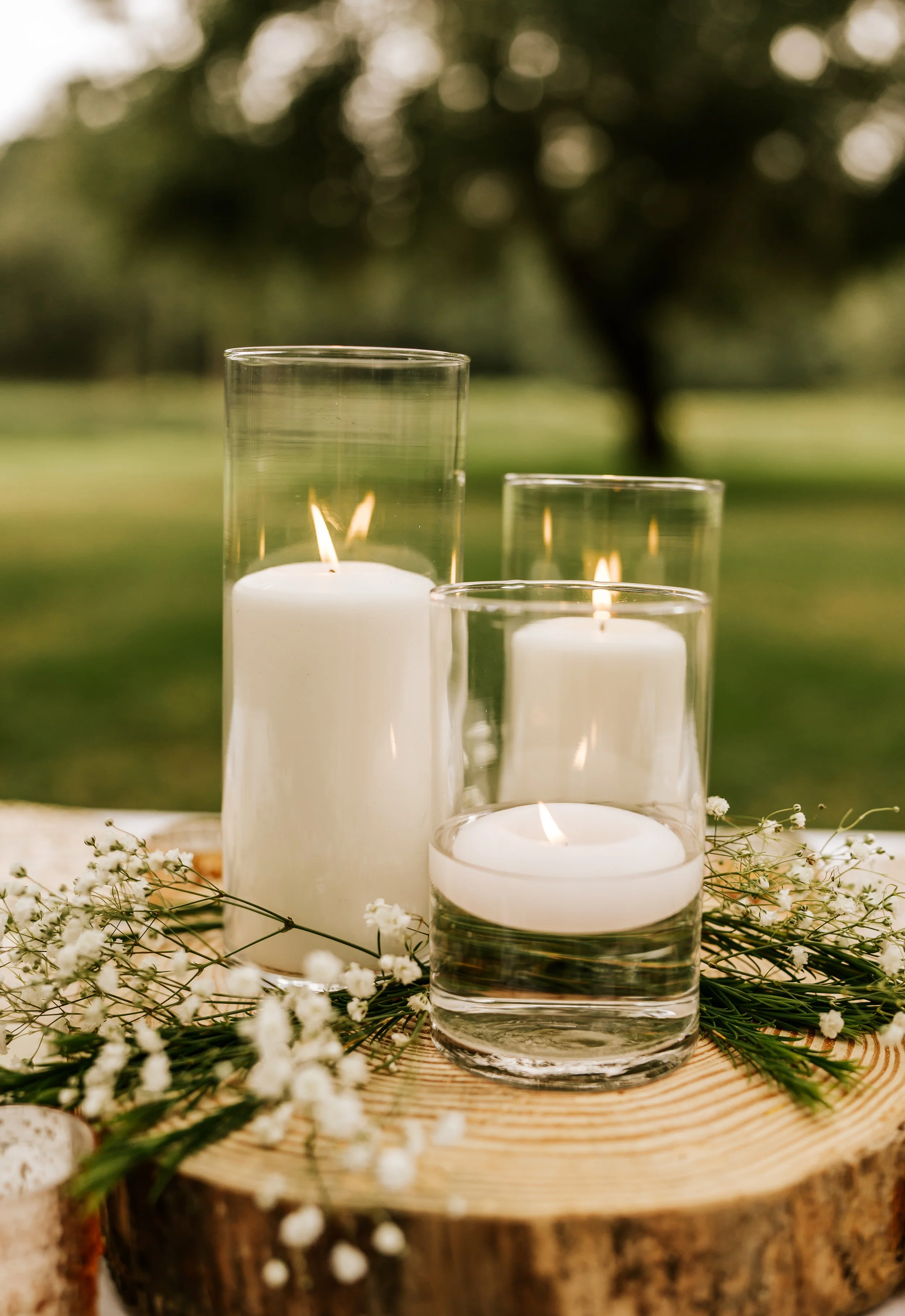 Three lit candles in glass holders arranged on a wooden surface, surrounded by small white flowers and greenery, outdoors with blurred trees in the background.