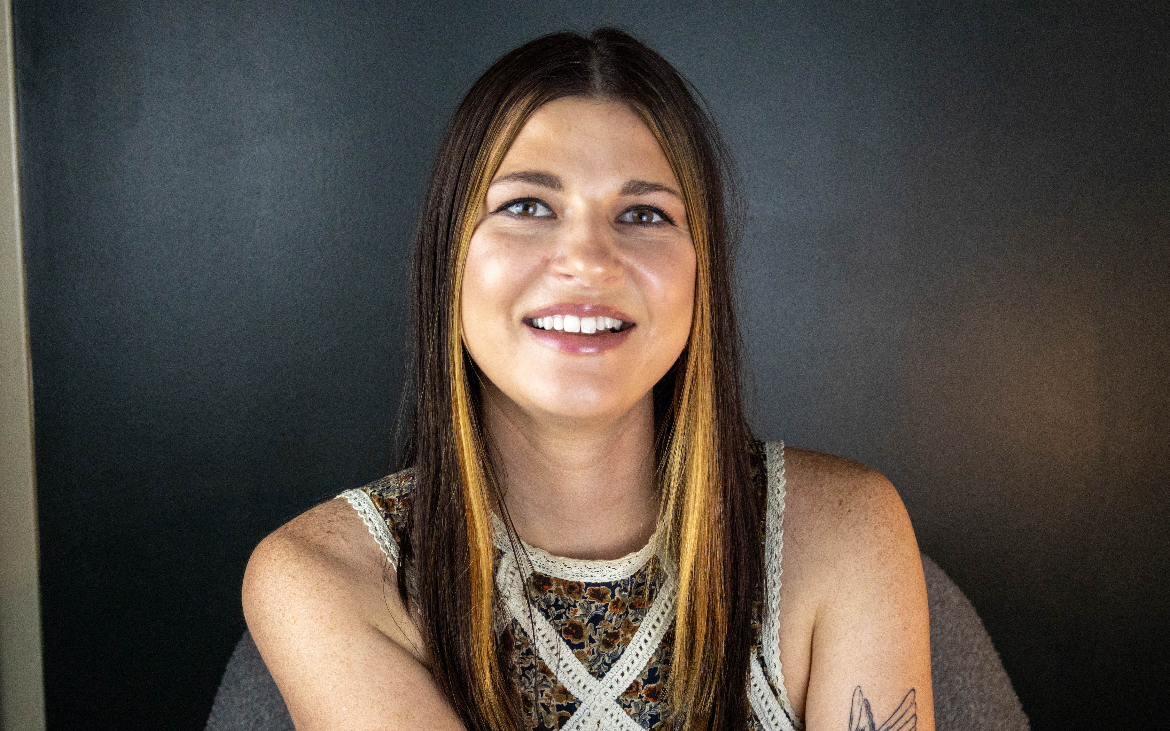 Smiling woman with long brown hair and a tattoo on her left arm, wearing a patterned sleeveless top, sitting against a dark background.
