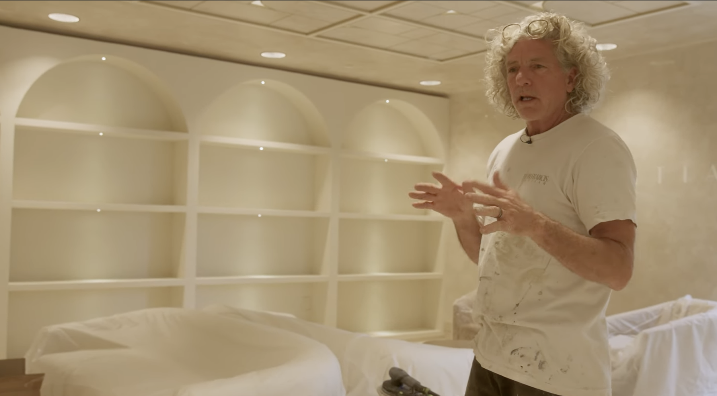 Man with curly gray hair explaining with hands in front of a cream-colored built-in shelving unit.
