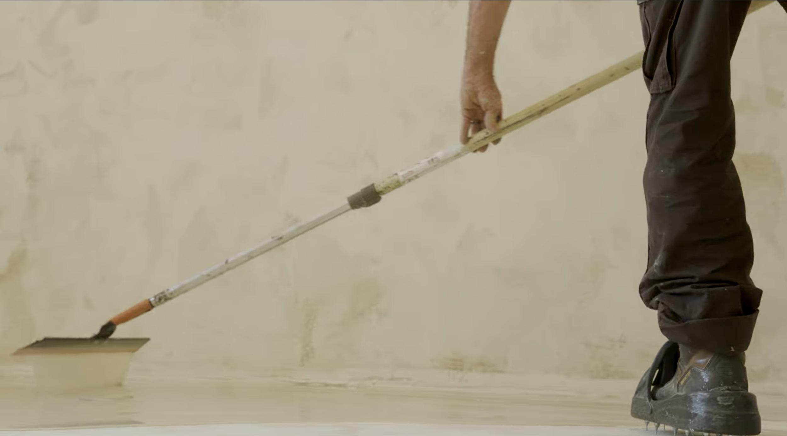 A person with brown work boots is using a drywall taper tool to smooth freshly applied joint compound on a wall.