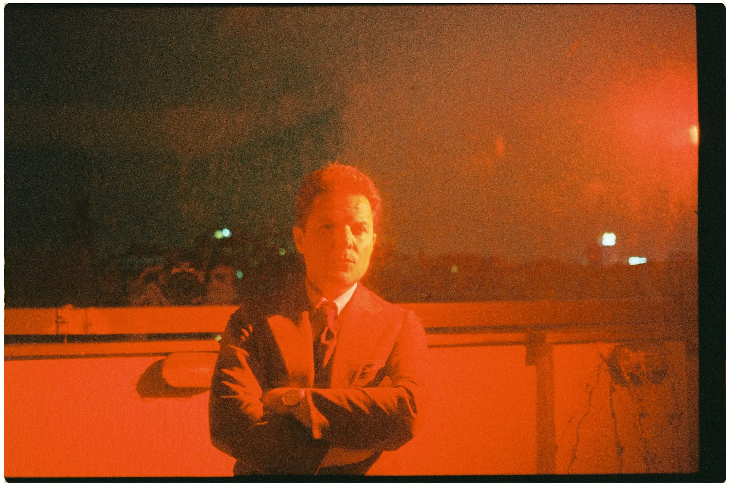 Mann in Anzug mit verschränkten Armen vor einer roten beleuchteten Wand, im Hintergrund drohne und Fotografen