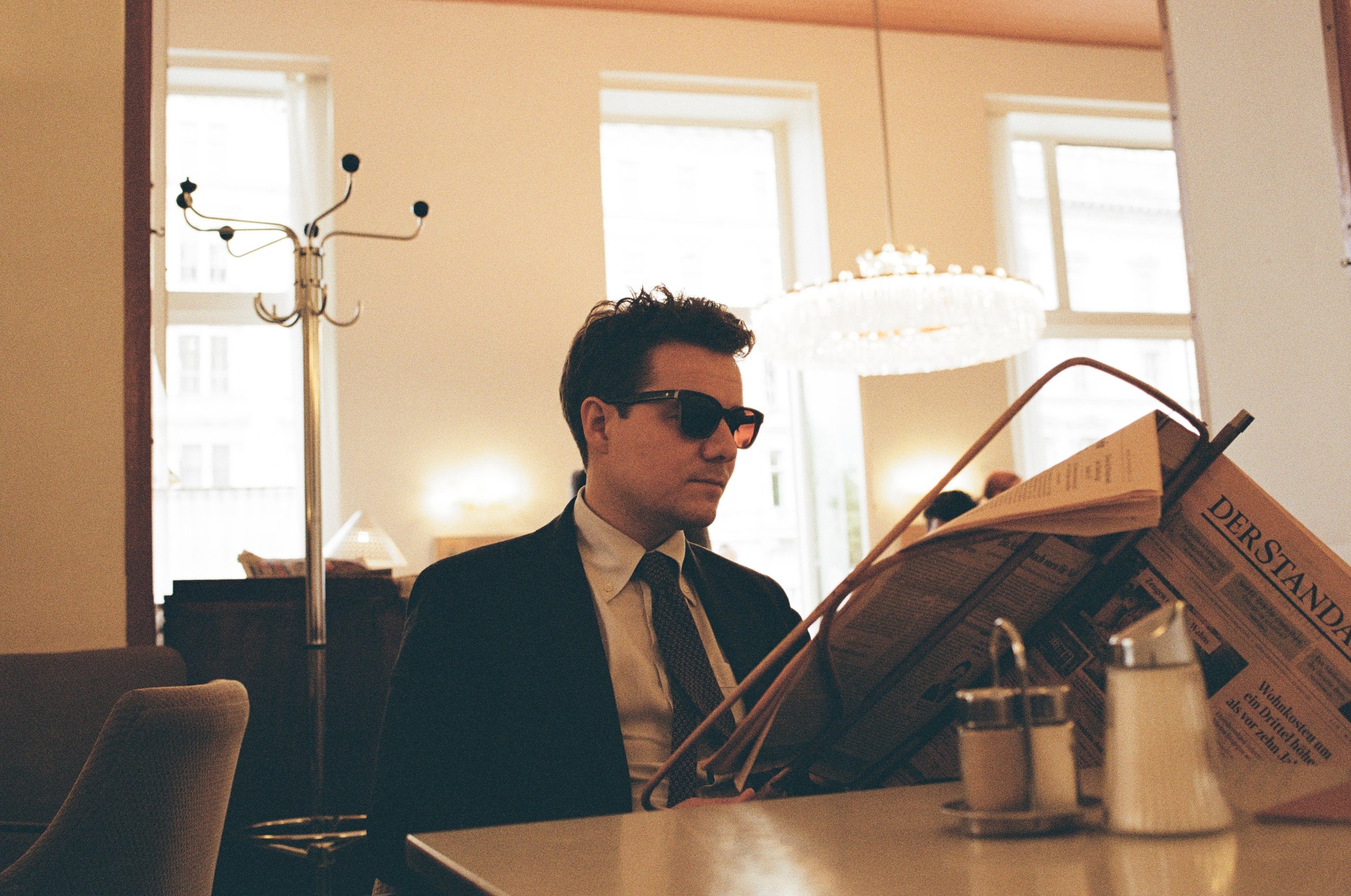 Junge Mann in Anzug und Sonnenbrille liest Zeitung in Café, im Hintergrund Kronleuchter und Fenster.
