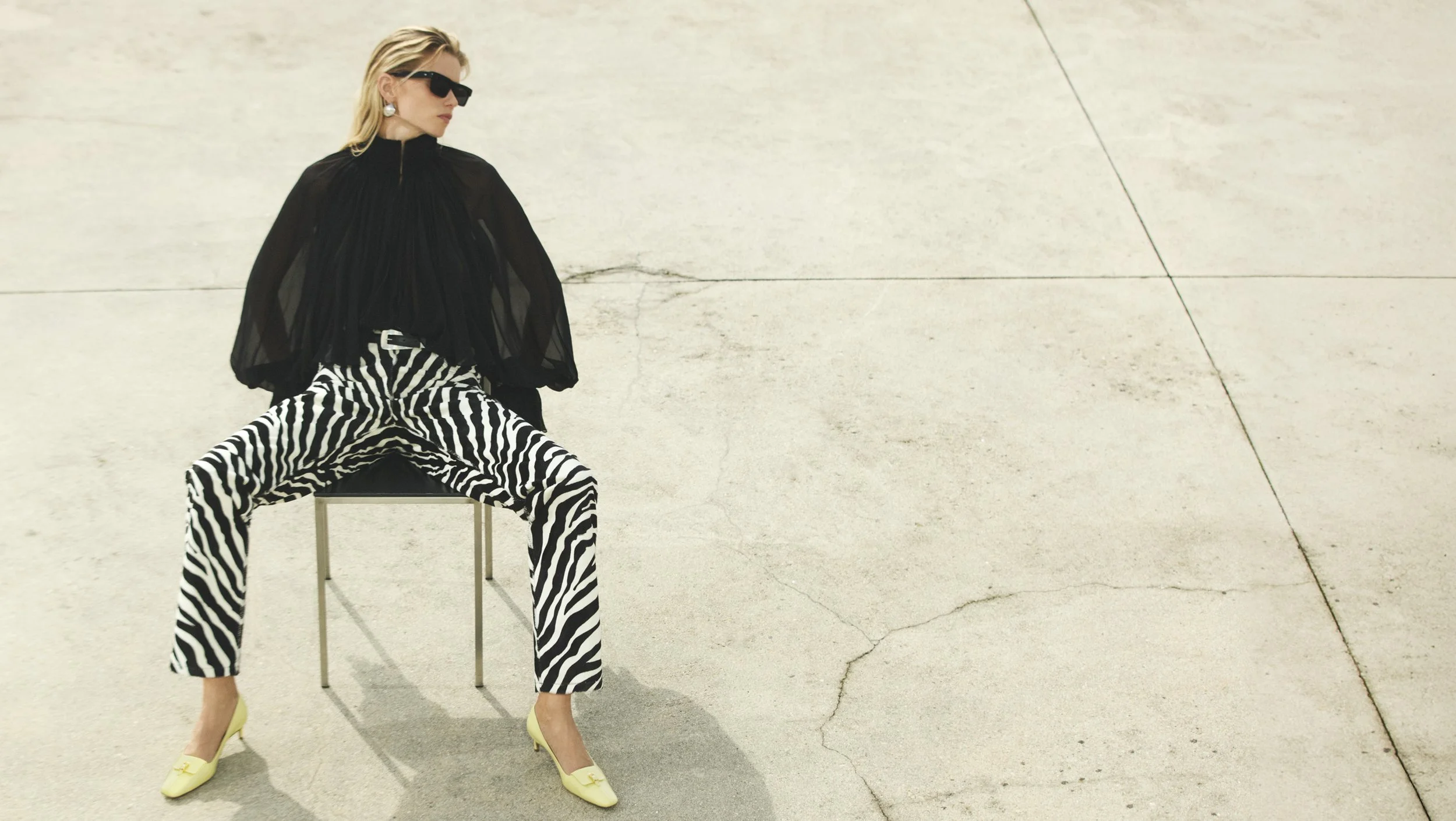 Model Abbey Lee with blonde hair, wearing black sunglasses, and black earrings, seated on a black chair, wearing a black sheer blouse, zebra print pants, and yellow high heels, poses against a plain, cracked concrete background. 