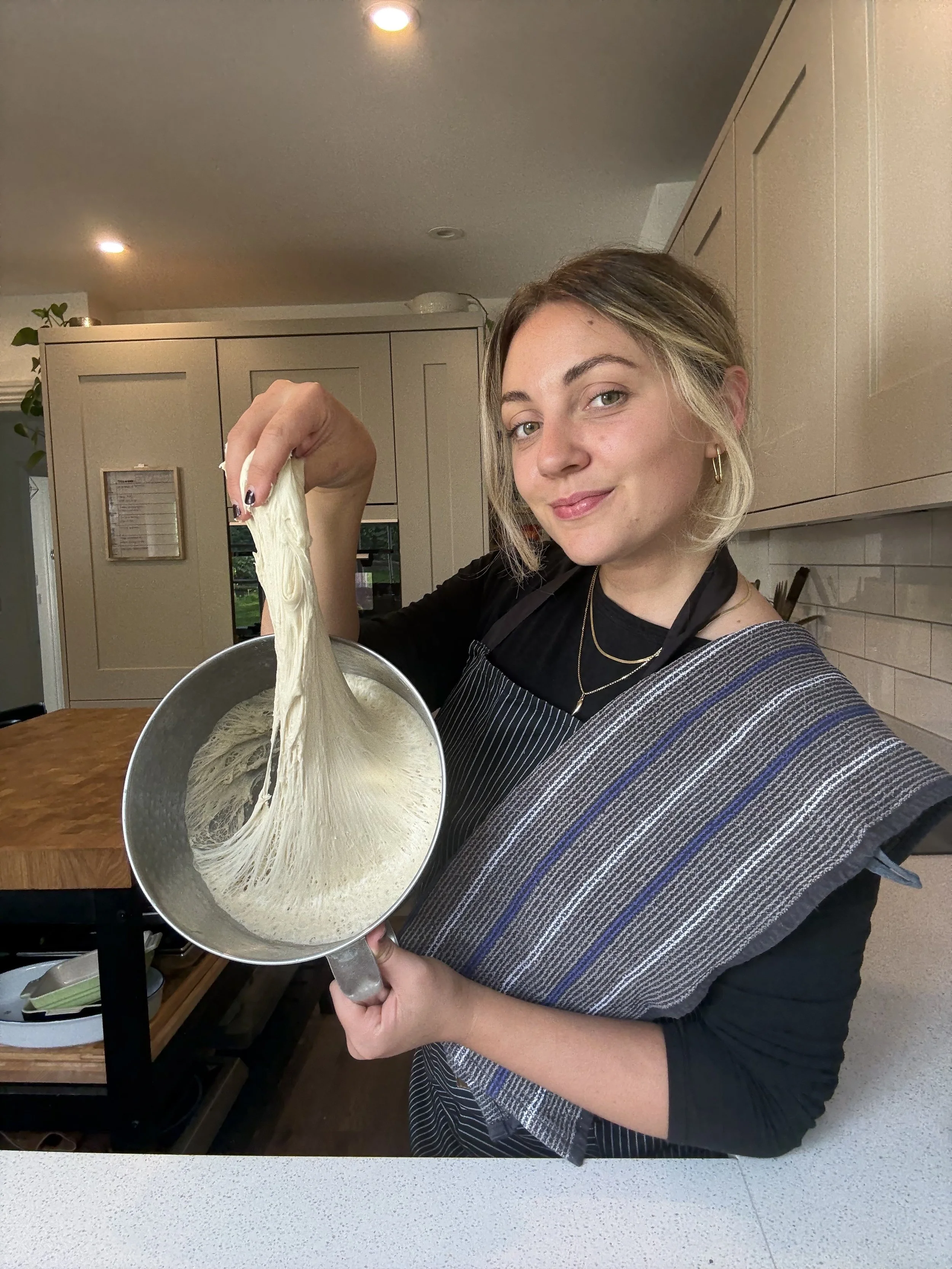 Woman holds a saucepan with stretchy dough, pulling it up to show its texture in a modern kitchen.