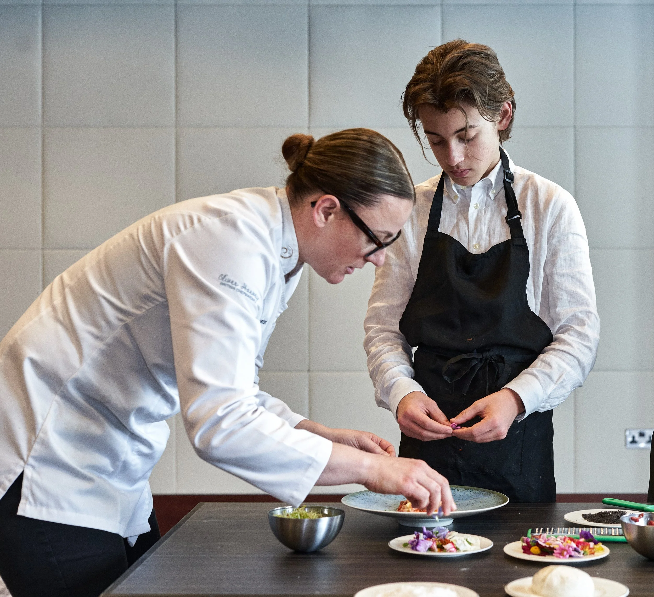 Two chefs, a male and a female, preparing food in a kitchen. The male is wearing glasses and a white chef's coat, and the female is wearing a white shirt with a black apron. They are working together at a black countertop with various colorful decorated cookies and ingredients displayed.