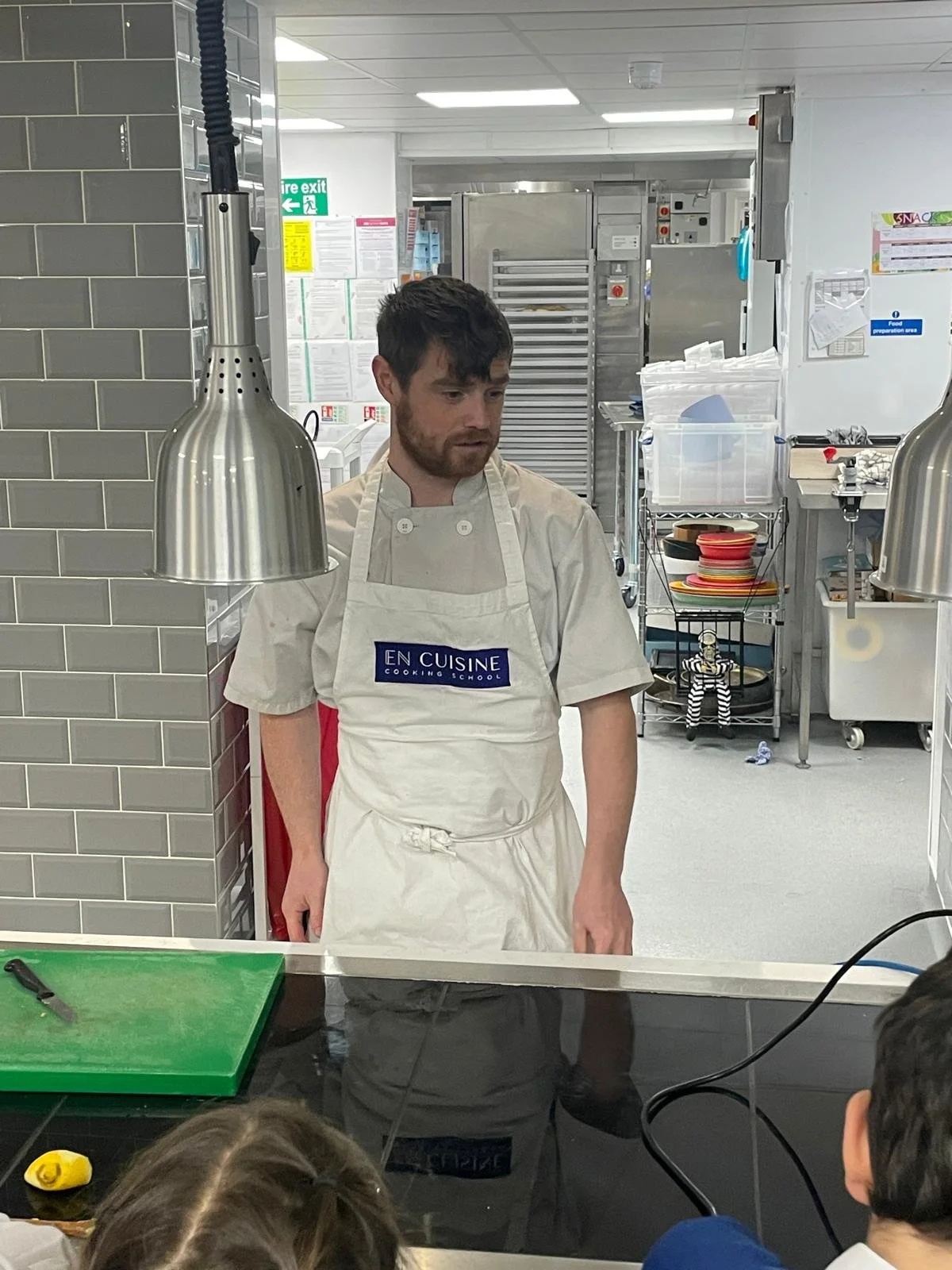 A chef wearing a white apron with 'EN CUISINE' written on it is standing behind a kitchen counter, looking down, with a teaching or demonstration environment in the background.