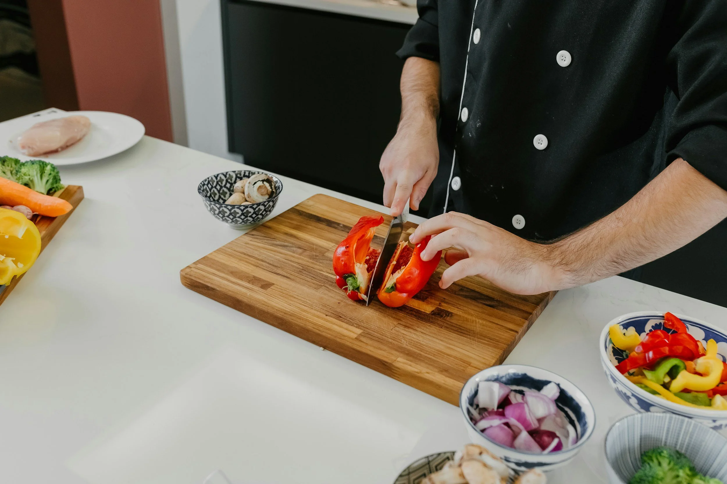 Person in black chef's coat slicing red bell peppers on a wooden cutting board in a kitchen with various vegetables around.