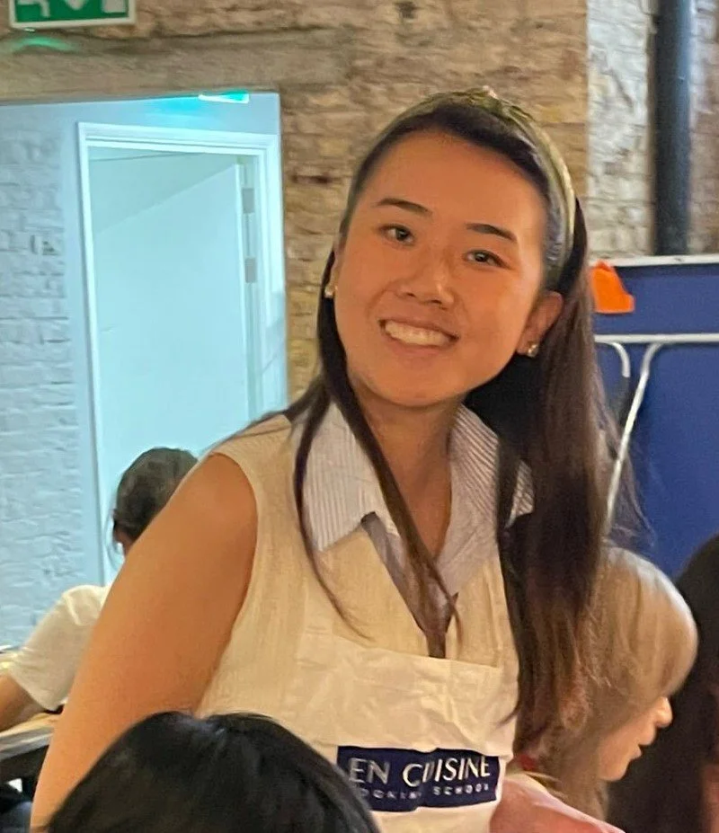 A smiling woman in a restaurant, wearing a sleeveless top and an apron, with other diners and brick walls visible in the background.