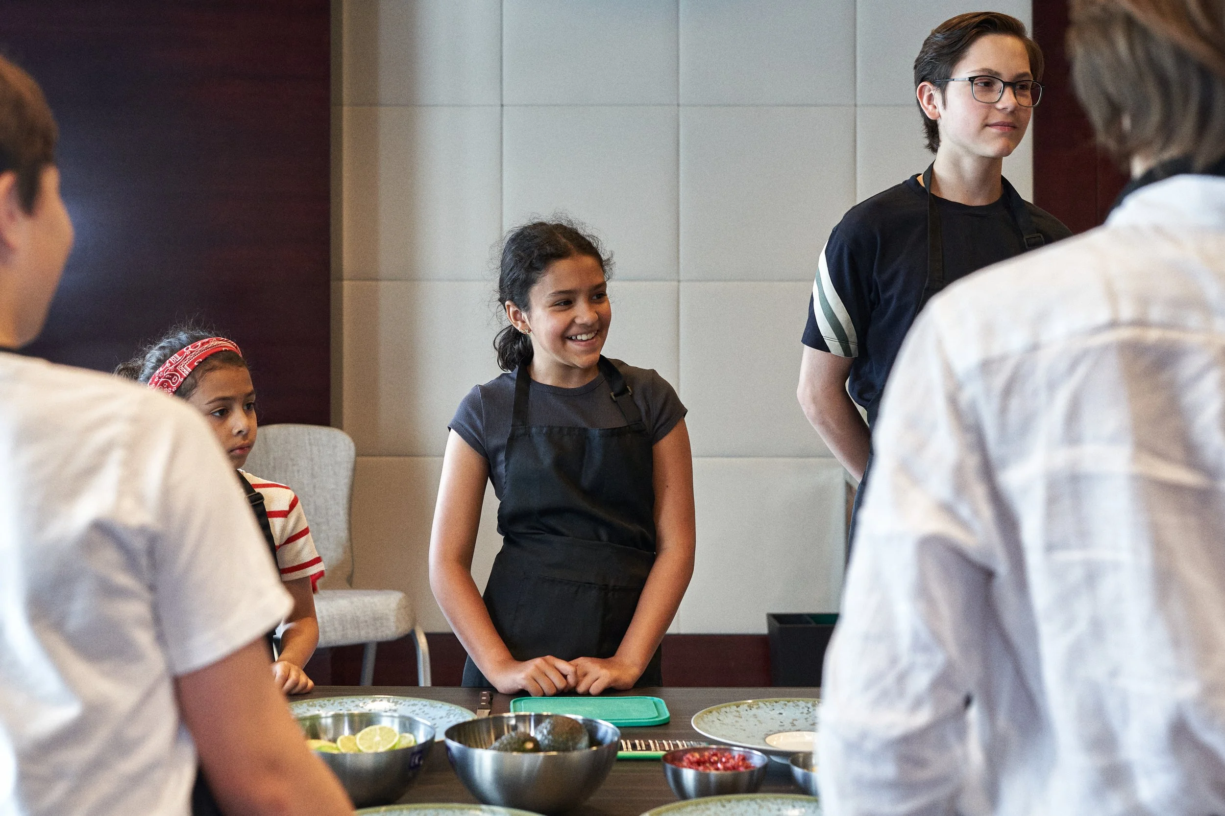 A group of children and a young adult standing around a table with bowls of ingredients. The young adult in the center, wearing glasses and a black shirt, is smiling as the children listen attentively.