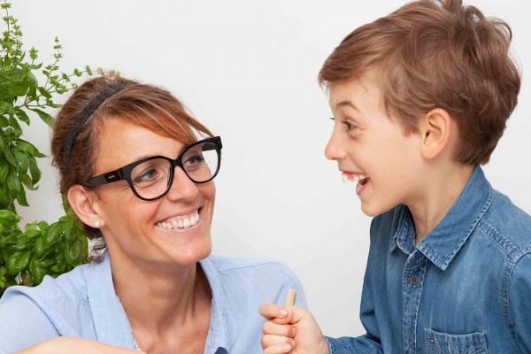 A woman with glasses and a headband smiling and looking at a smiling boy in a denim shirt, who is holding her hand.