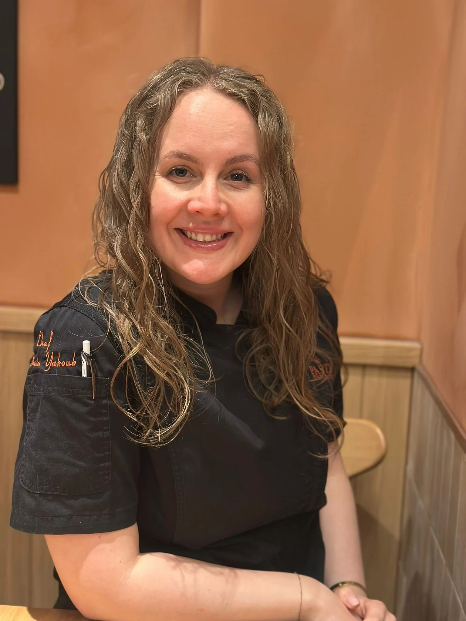 A woman with long curly brown hair wearing a black chef's coat, sitting in a restaurant or cafe with wooden walls, smiling at the camera.