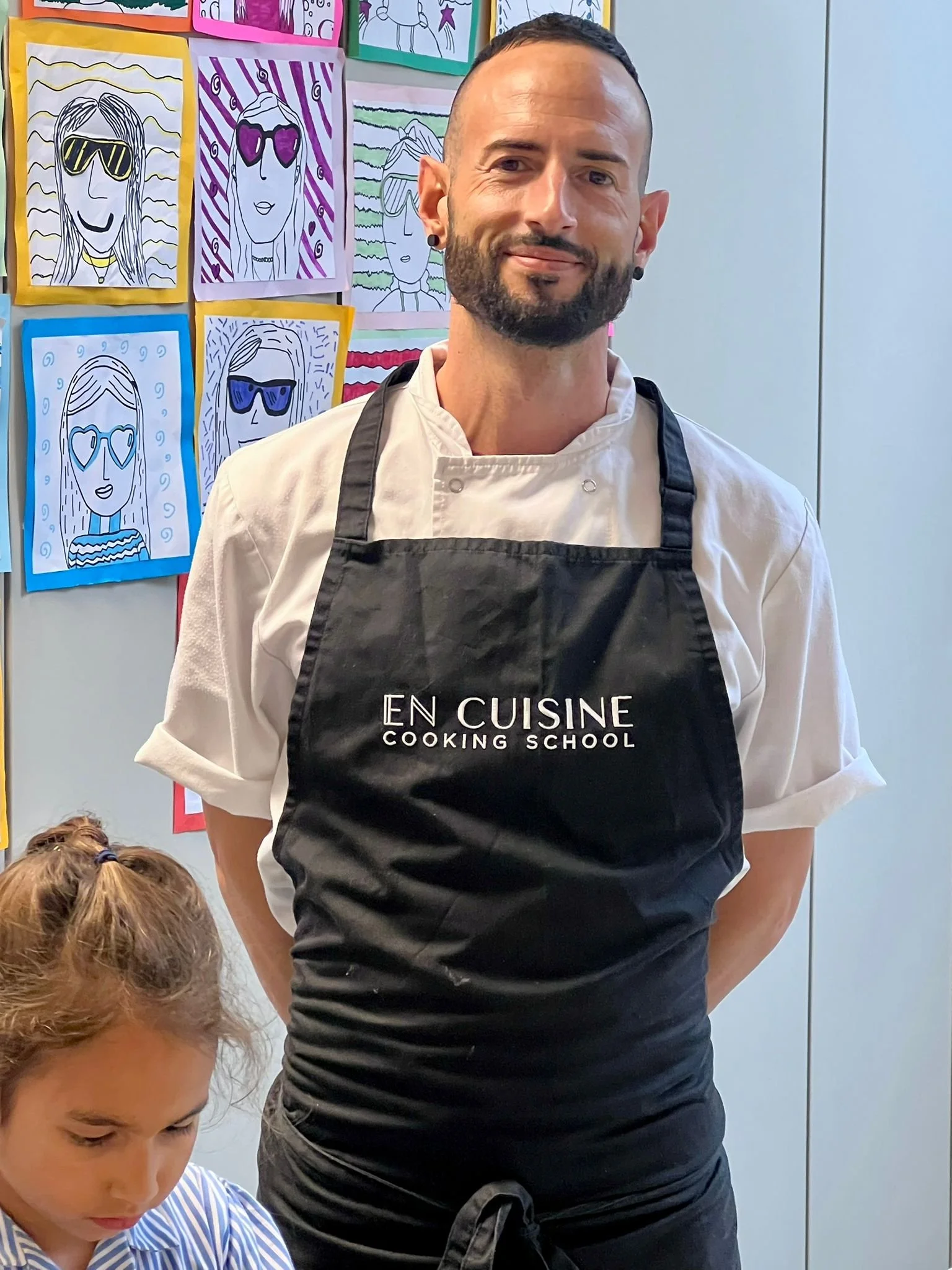 A man wearing a white chef's coat and a black apron with the logo 'En Cuisine Cooking School', standing with a woman or girl with curly hair in front of colorful drawings of women wearing sunglasses displayed on a wall.