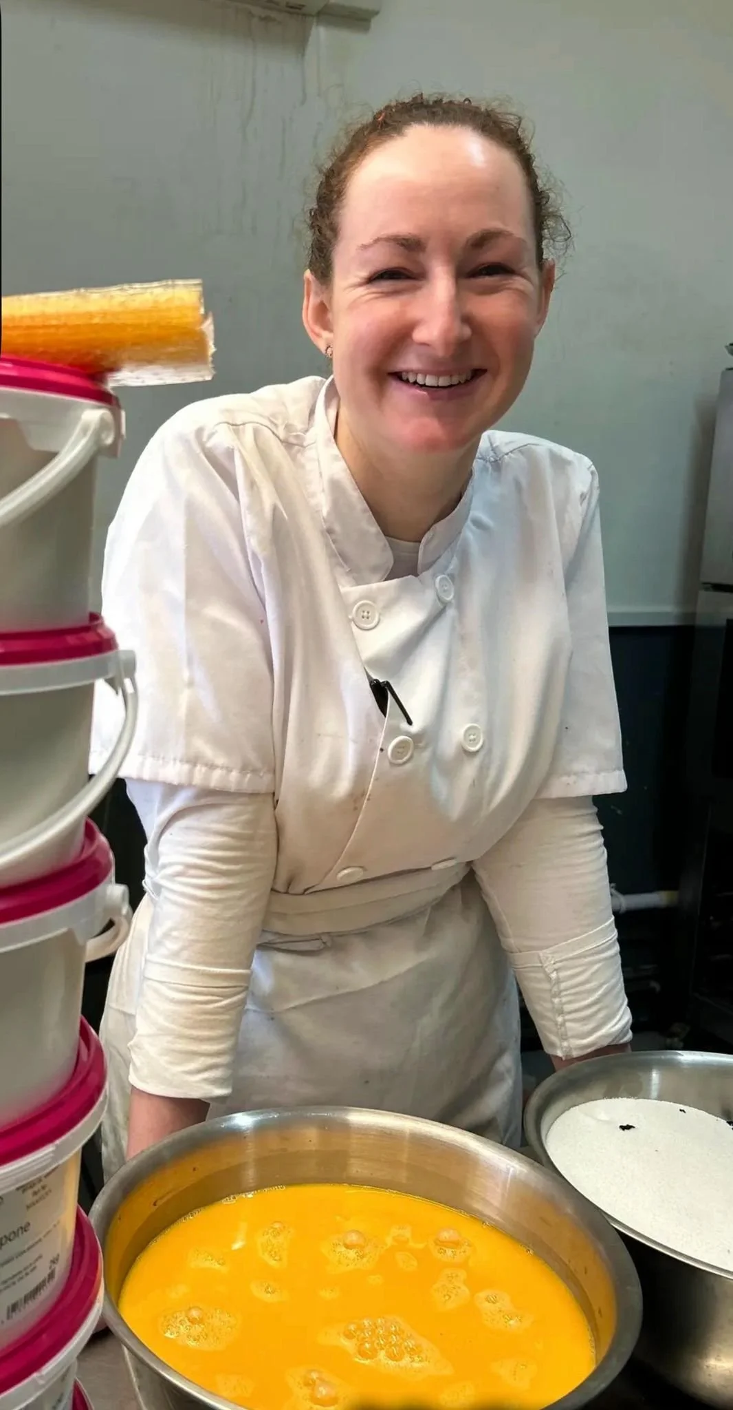 A woman in a chef's coat smiling at the camera, with bowls of beaten eggs and flour in front of her in a kitchen.