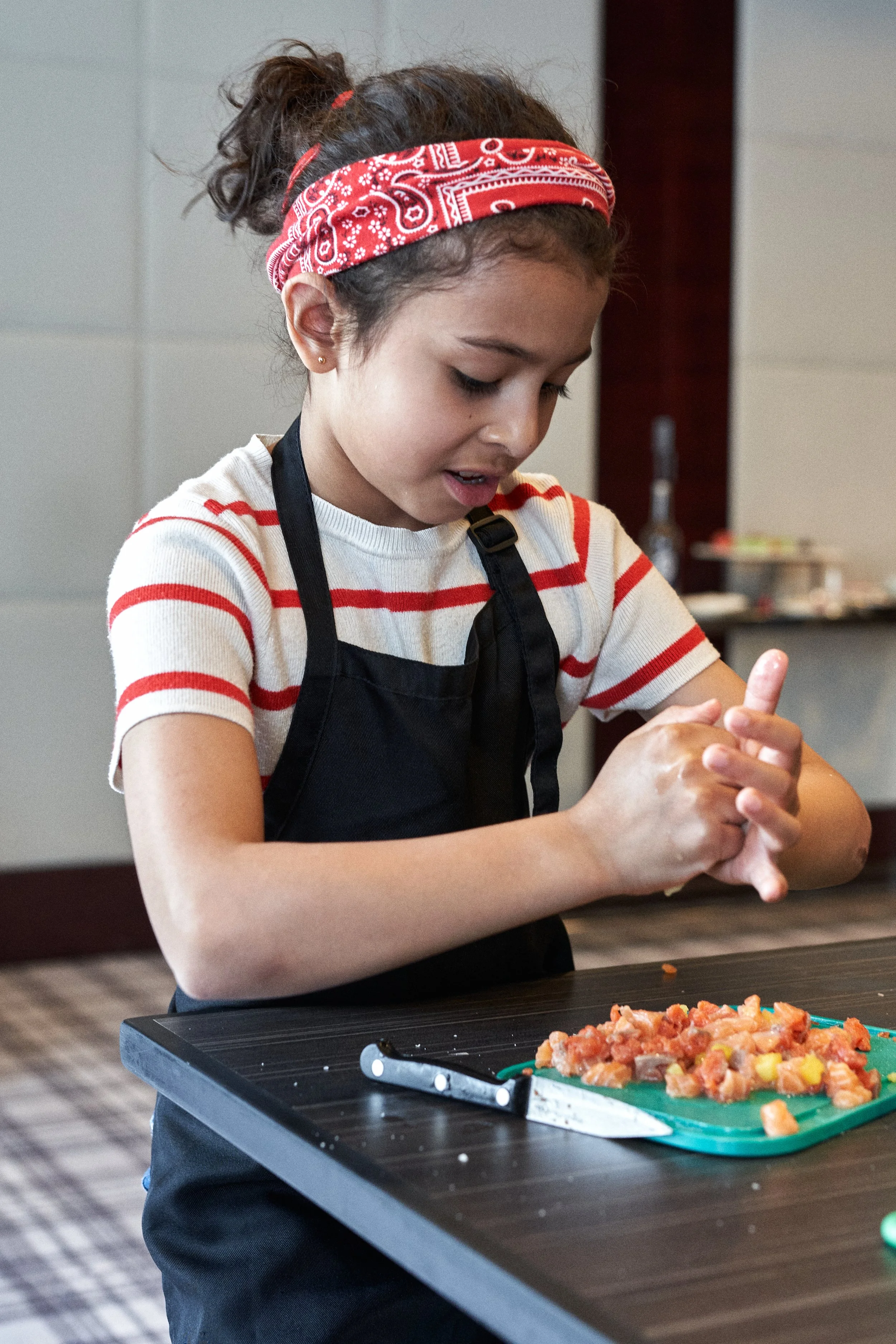 A young girl with dark hair tied in a ponytail, wearing a red bandana and a striped shirt, preparing diced vegetables or meat on a blue cutting board in a kitchen. She is wearing a black apron and is focused on her task.