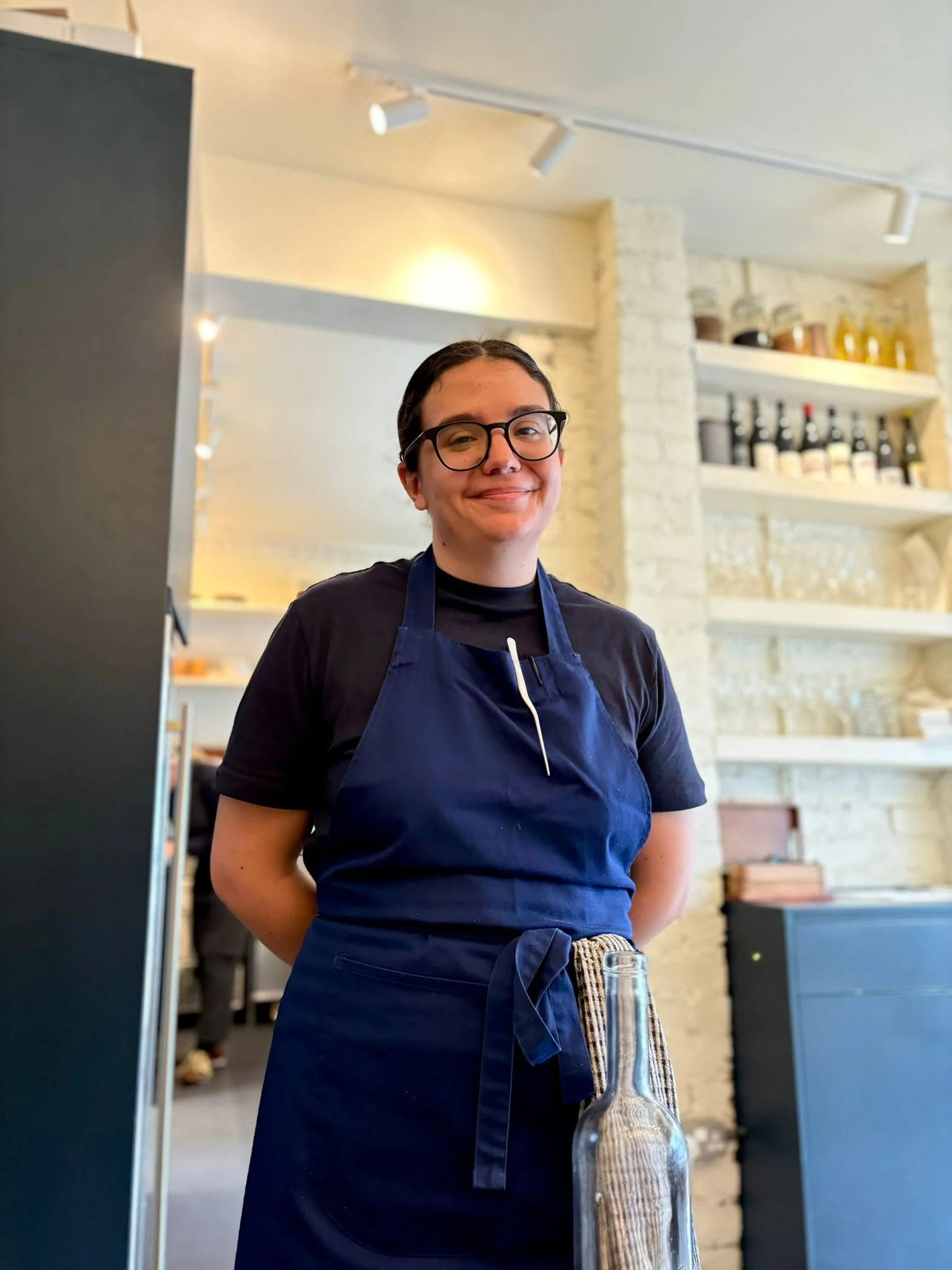 A smiling woman wearing glasses and a navy apron stands in a modern kitchen or café setting with shelves of bottles and jars behind her, and a glass bottle on the table in front of her.