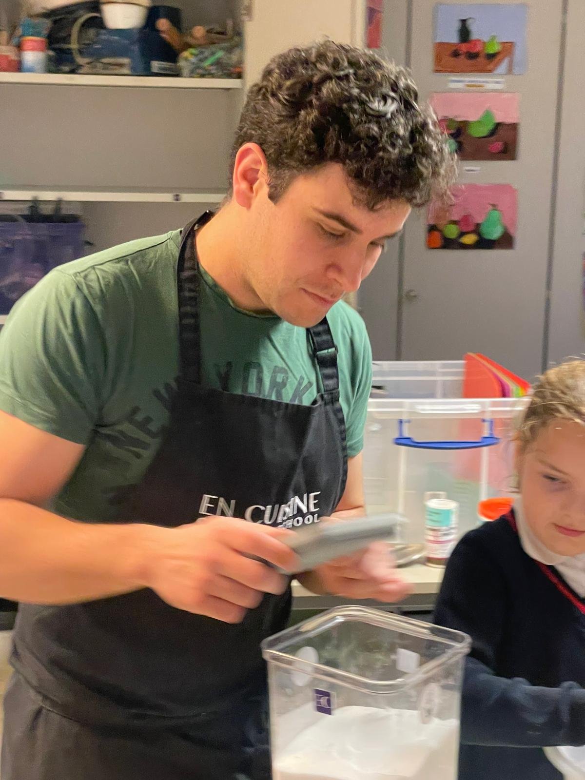 A young man with curly dark hair wearing a green t-shirt and a black apron with white text, in a kitchen setting, looking down at a device in his hands. Next to him, a young girl with blonde hair and a dark sweater is partially visible. The background includes shelves and containers.