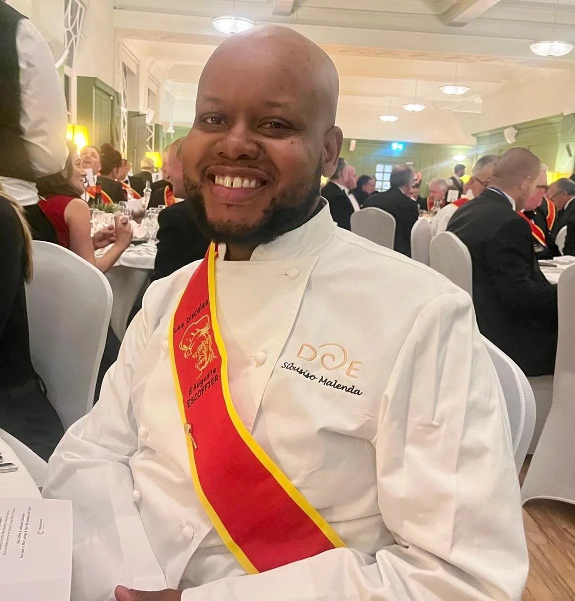 A smiling chef in a white uniform with a red and yellow sash at a formal event with many people in formal attire, dining and socializing in the background.