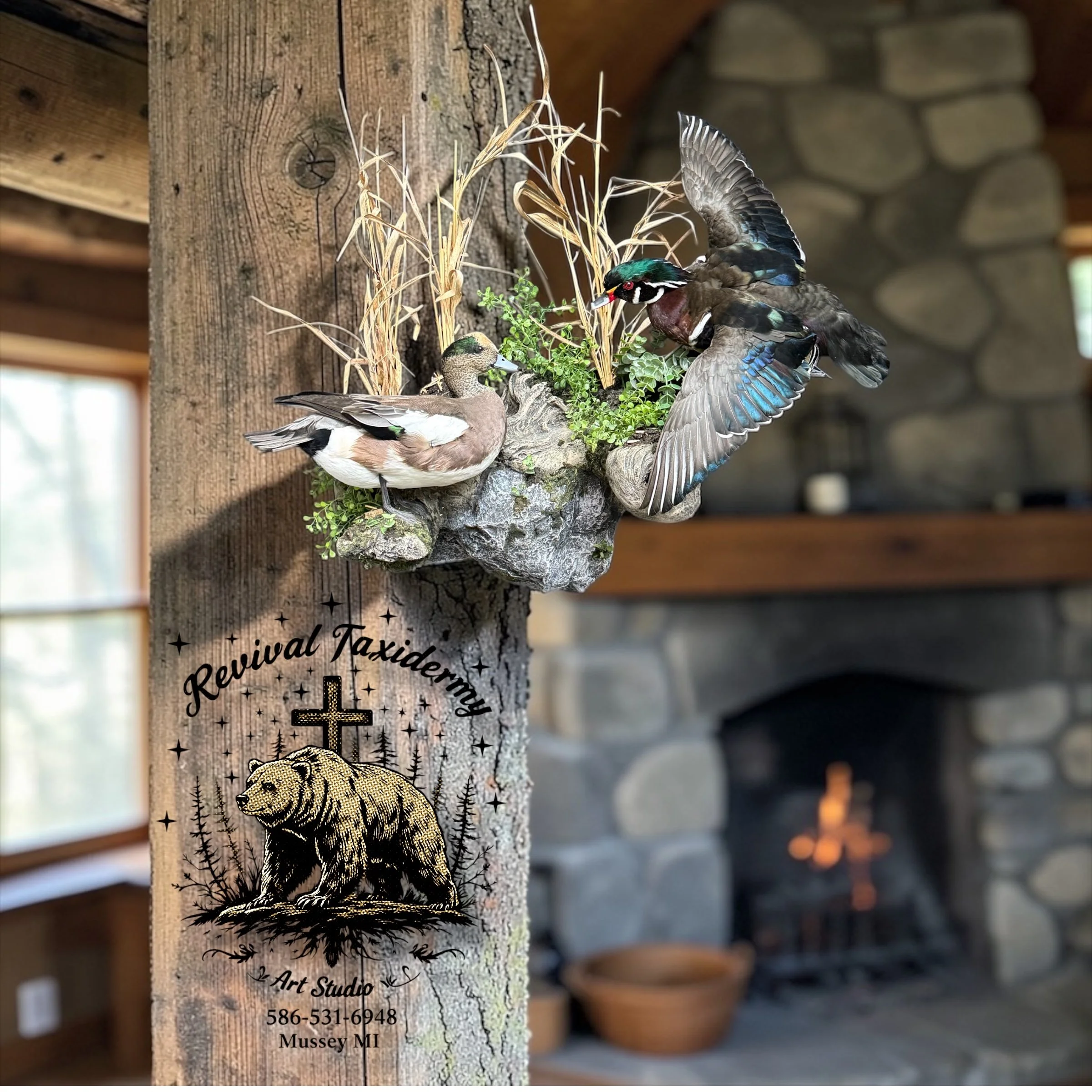 Decorative wall art of a bear, cross, and trees with text 'Revival Taxidermy' and art studio contact information, mounted on a wooden post with mounted ducks and wood plants, in front of a stone fireplace.
