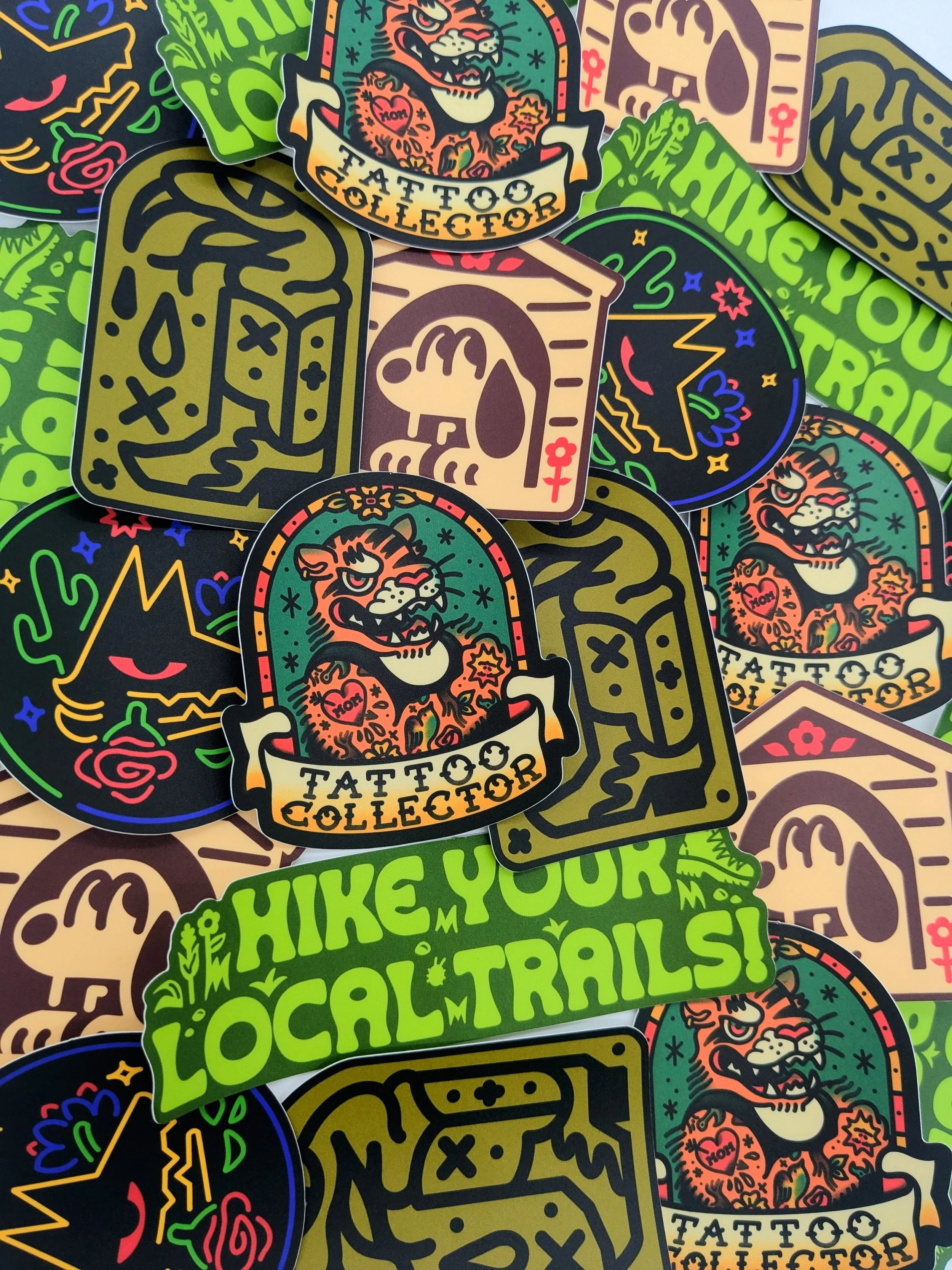 Various vinyl stickers in a pile. It includes: a Tattoo Collector, Doghouse, Boot Snake, and Hike Your Local Trails sticker.