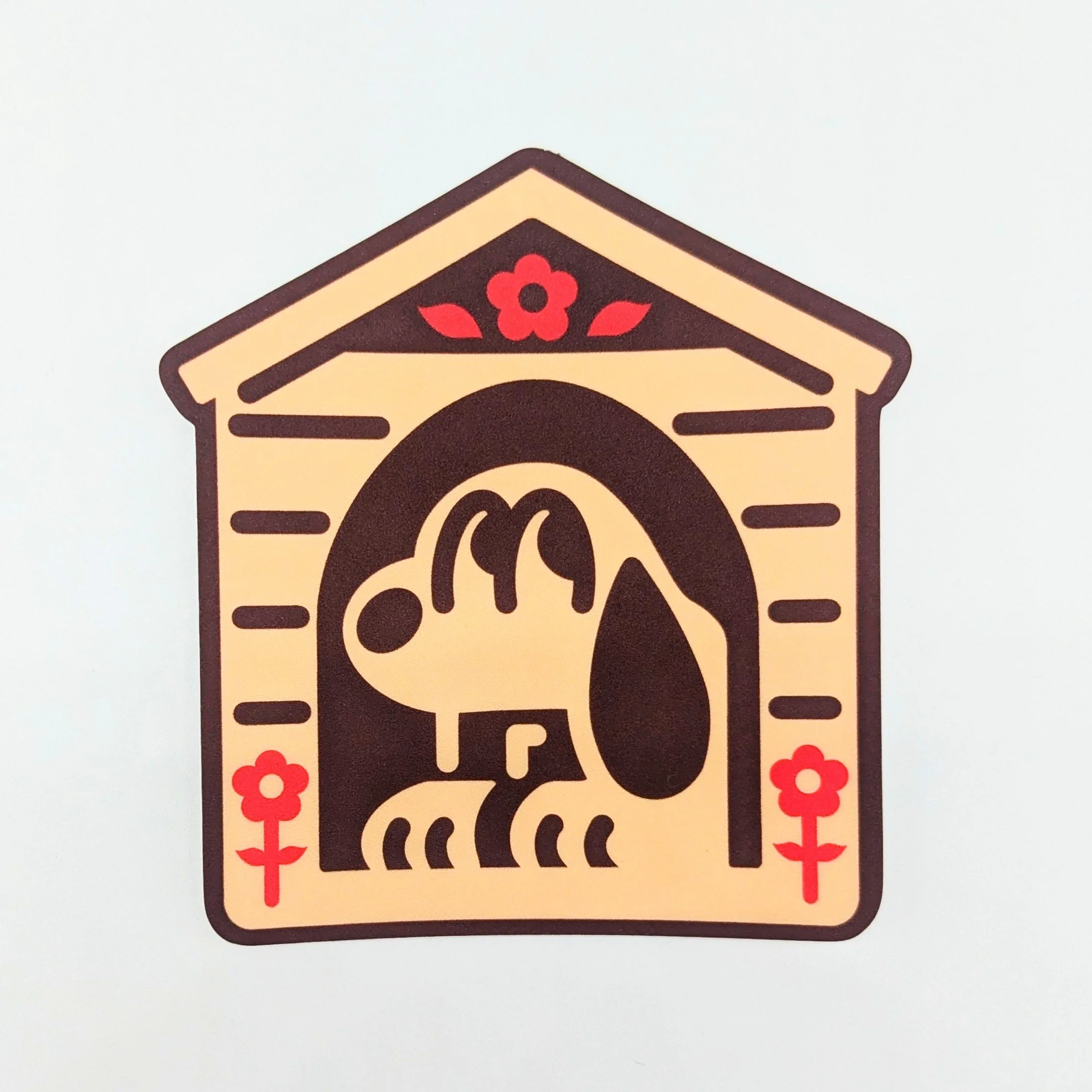 A vinyl sticker laying on a white backdrop. The sticker features a cartoon dog similar to Snoopy, sitting in a doghouse, with decorative red flowers. The sticker is called the Doghouse Sticker.