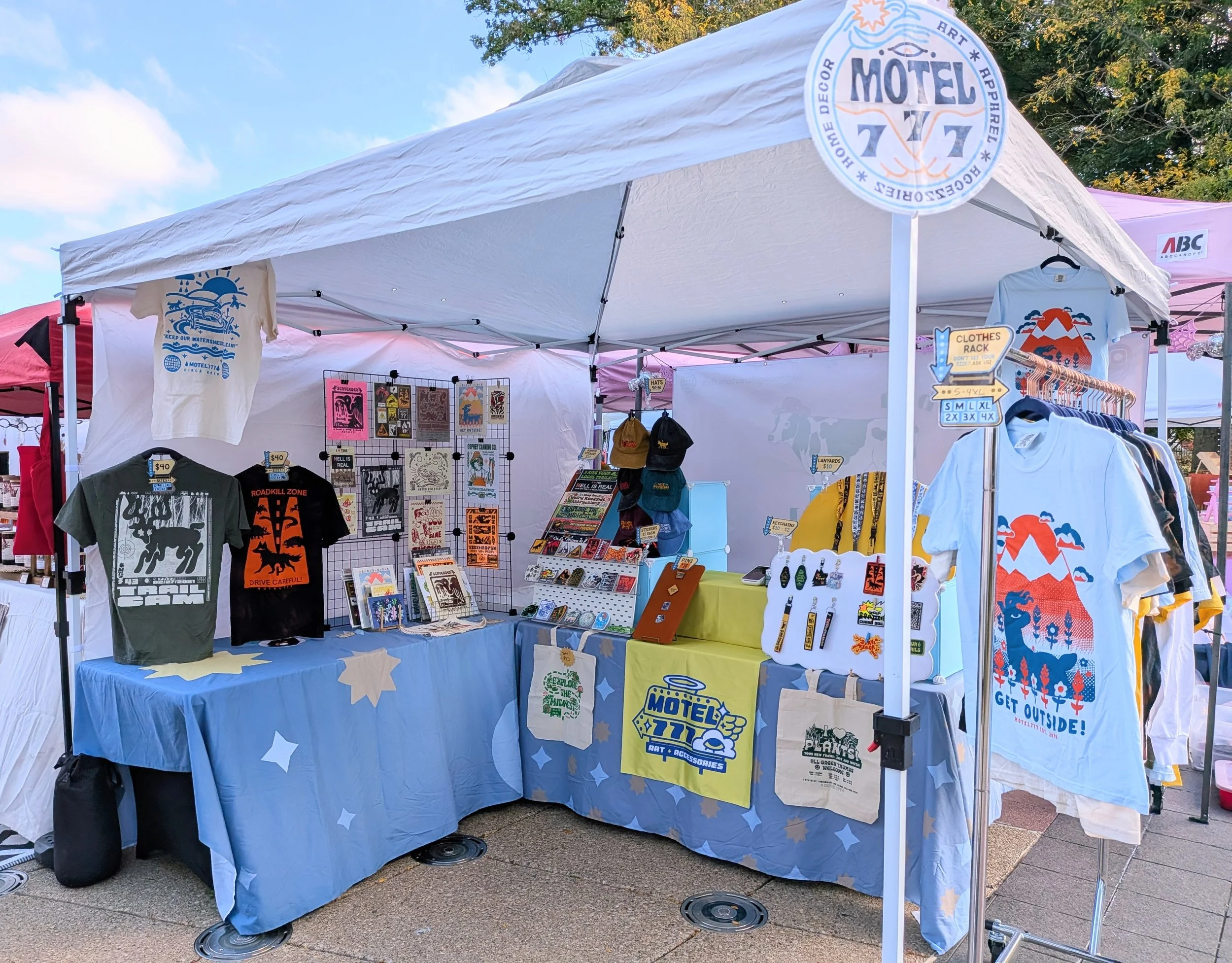 Outdoor set-up for 2026 City Flea craft market. It features a display of apparel, accessories & wall decor, all under a white tent canopy.