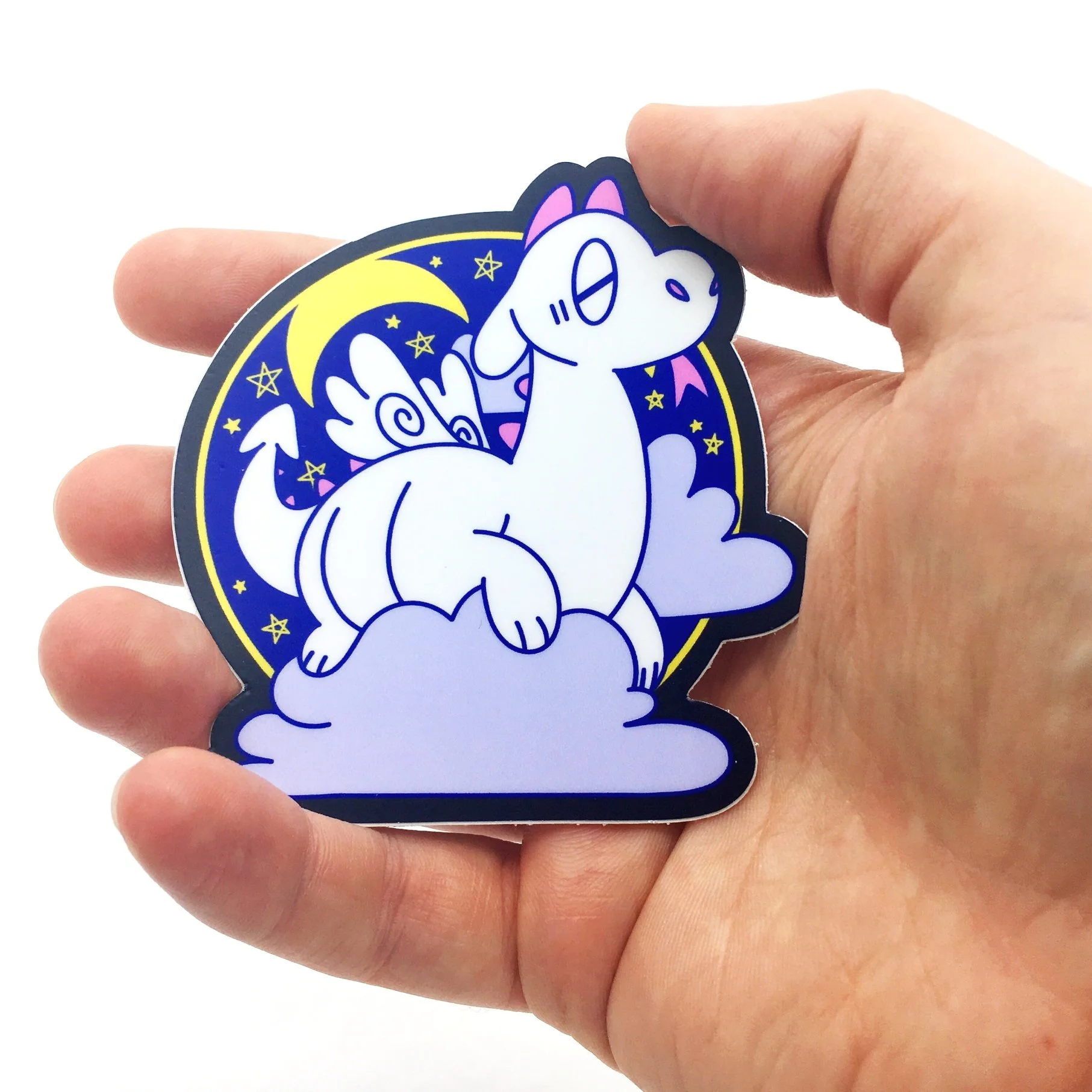 A hand holding a vinyl sticker against a white backdrop. The sticker features a sleeping cute white dragon on a cloud, surrounded by the moon and night sky. It is called the Dreamtime Dragon sticker.
