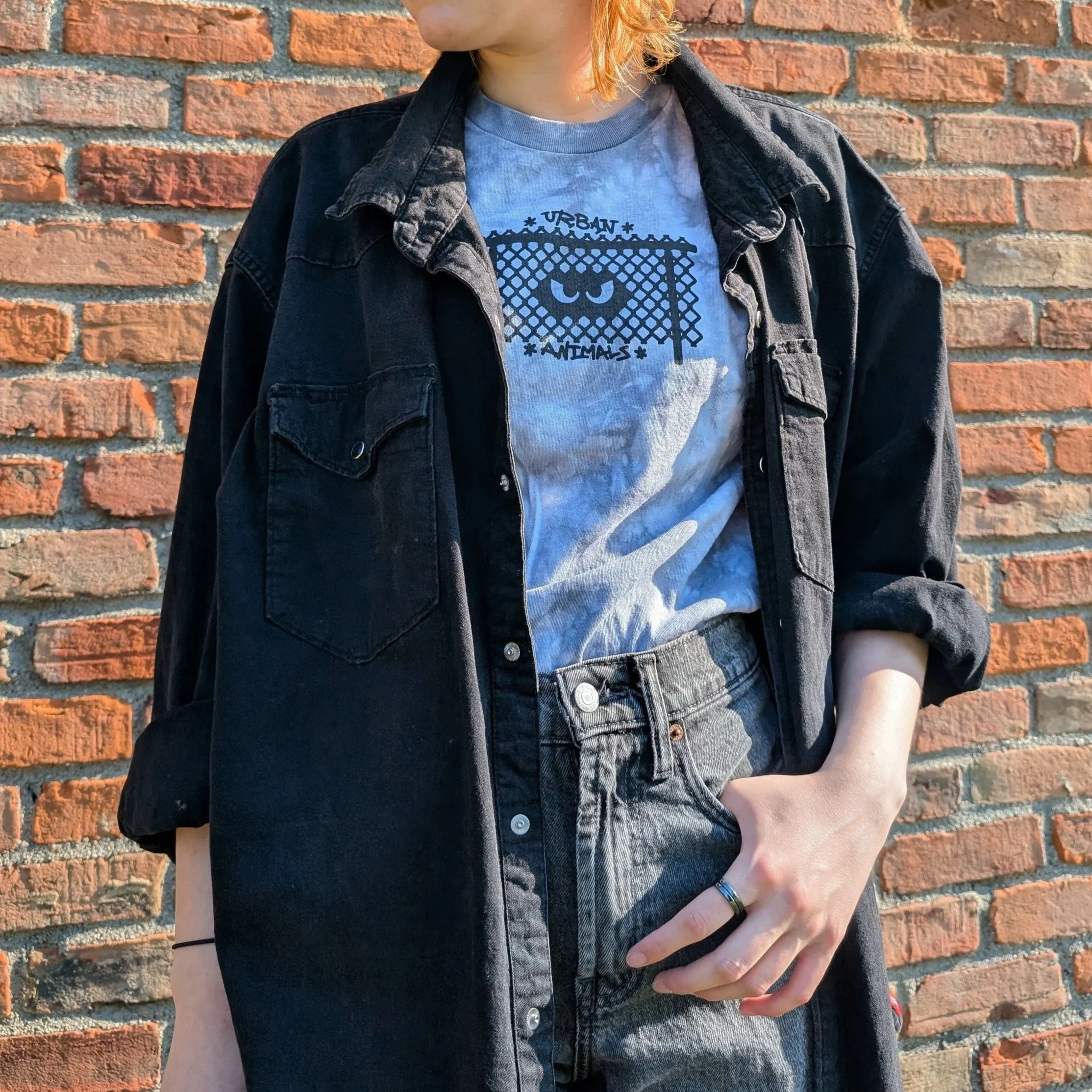 A person wearing a black denim jacket over an Urban Animals tee, standing against a red brick wall.