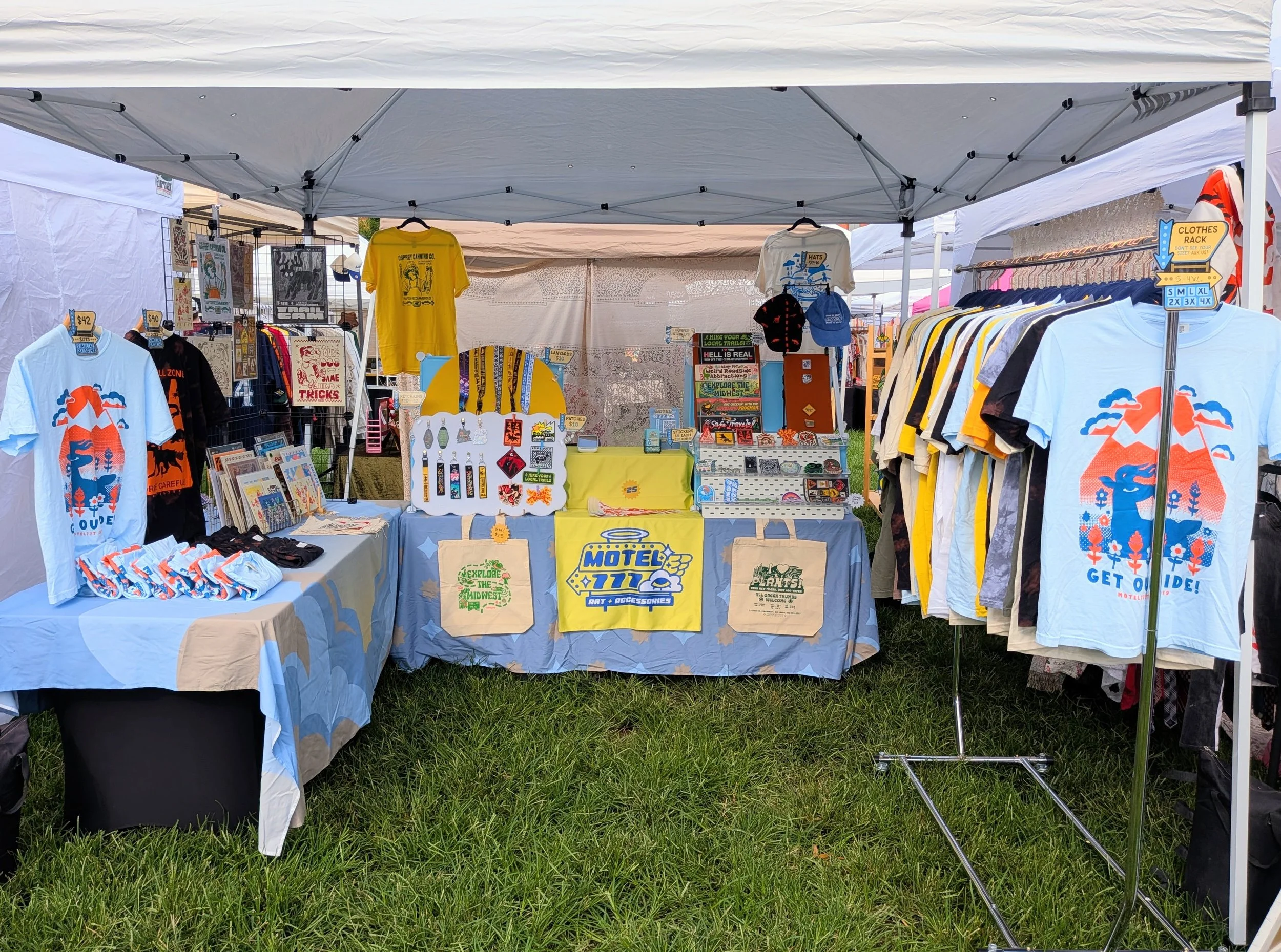 An outdoor booth at a market, with a table underneath a white canopy tent. It features apparel, accessories, and home decor.