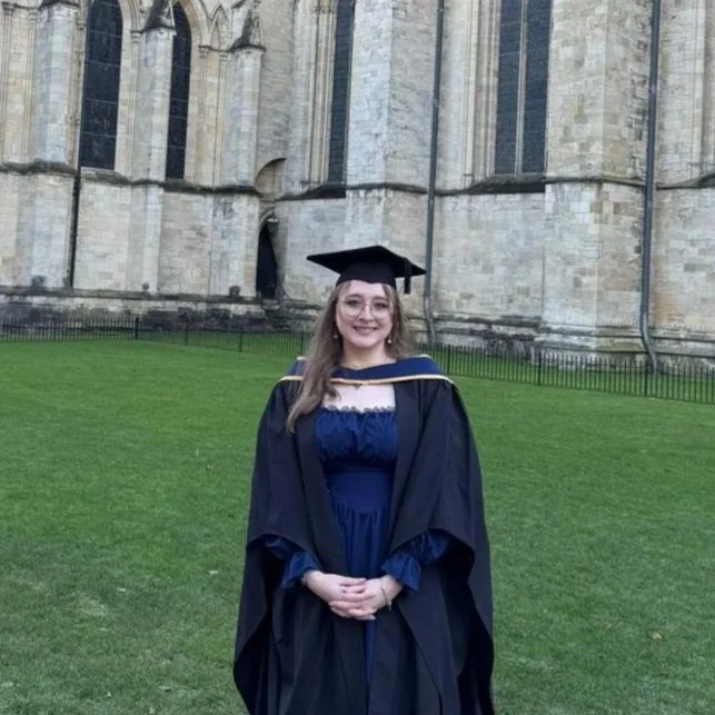 A young woman in graduation cap and gown standing outdoors on a grassy area in front of a historic stone building.