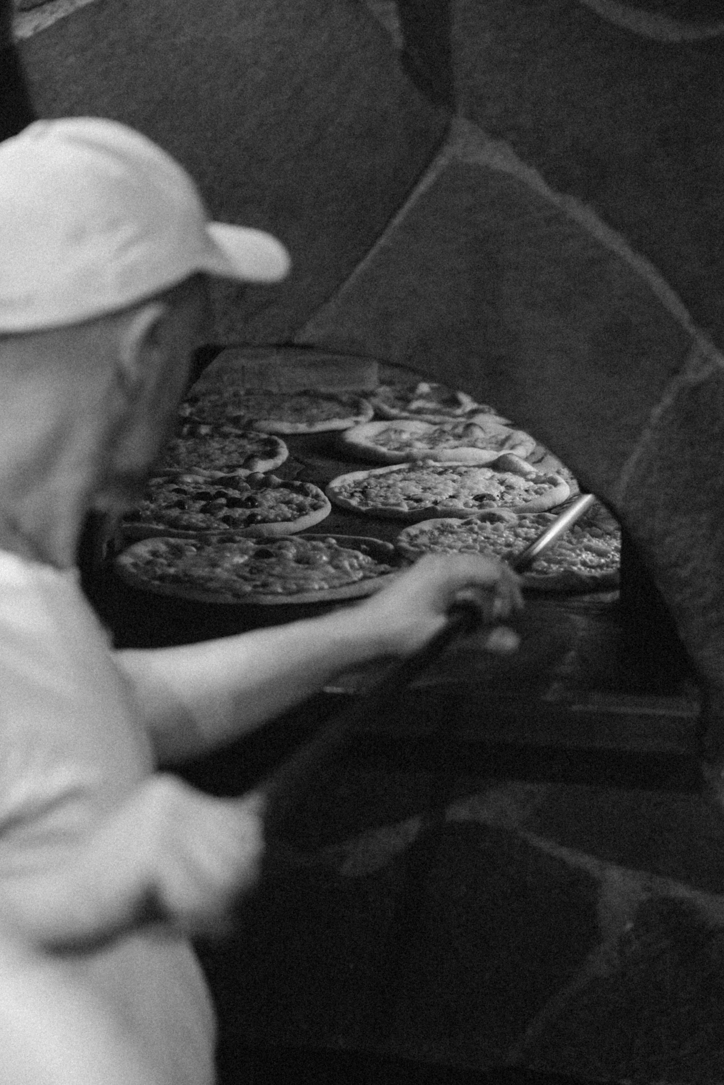 Eine Person, die Pizza backt, mit mehreren Pizzen auf einem Ofenrost. Die Person trägt eine weiße Schürze und eine weiße Mütze.
