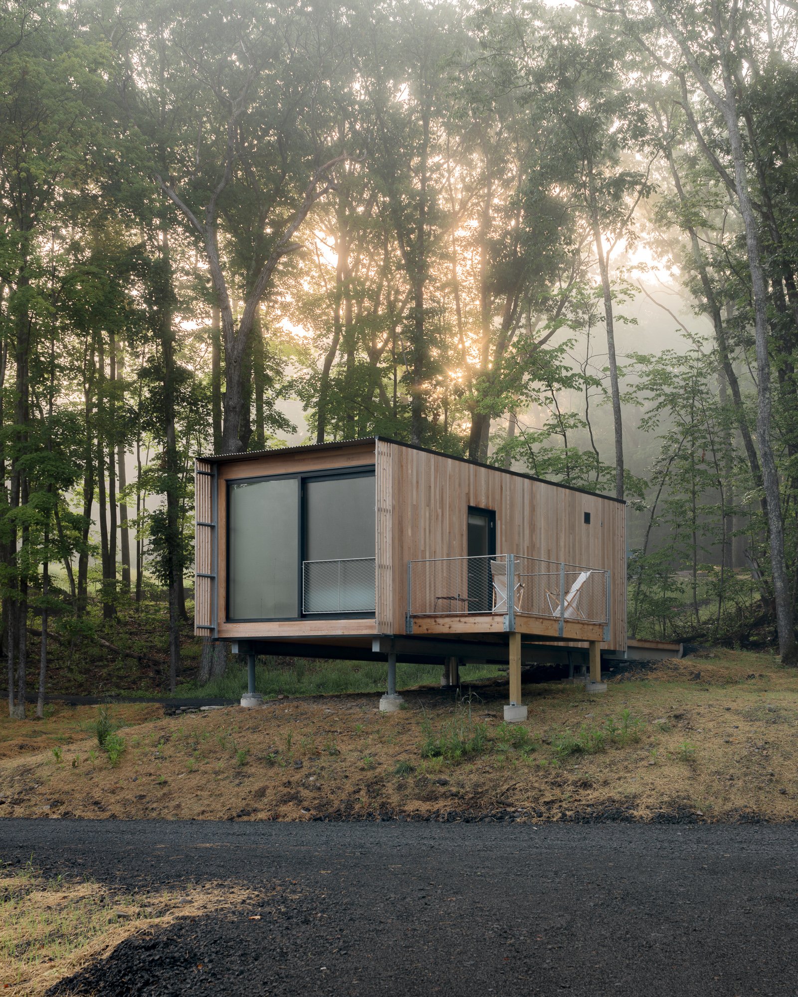 A small modern wooden house with a balcony and glass sliding door, situated in a forest clearing during sunrise or sunset.