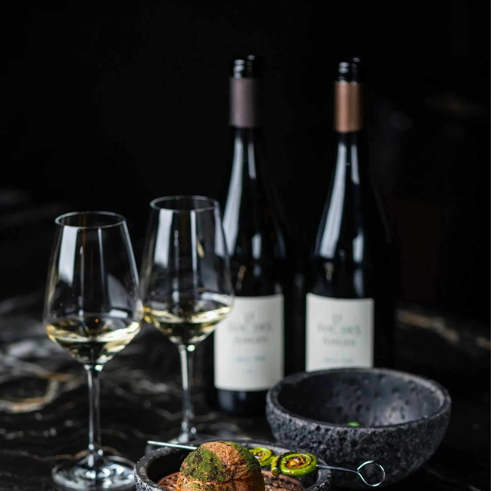 Two bottles of white wine with two poured glasses and a course on the black marble table at Class Act, for the Showcase membership.
