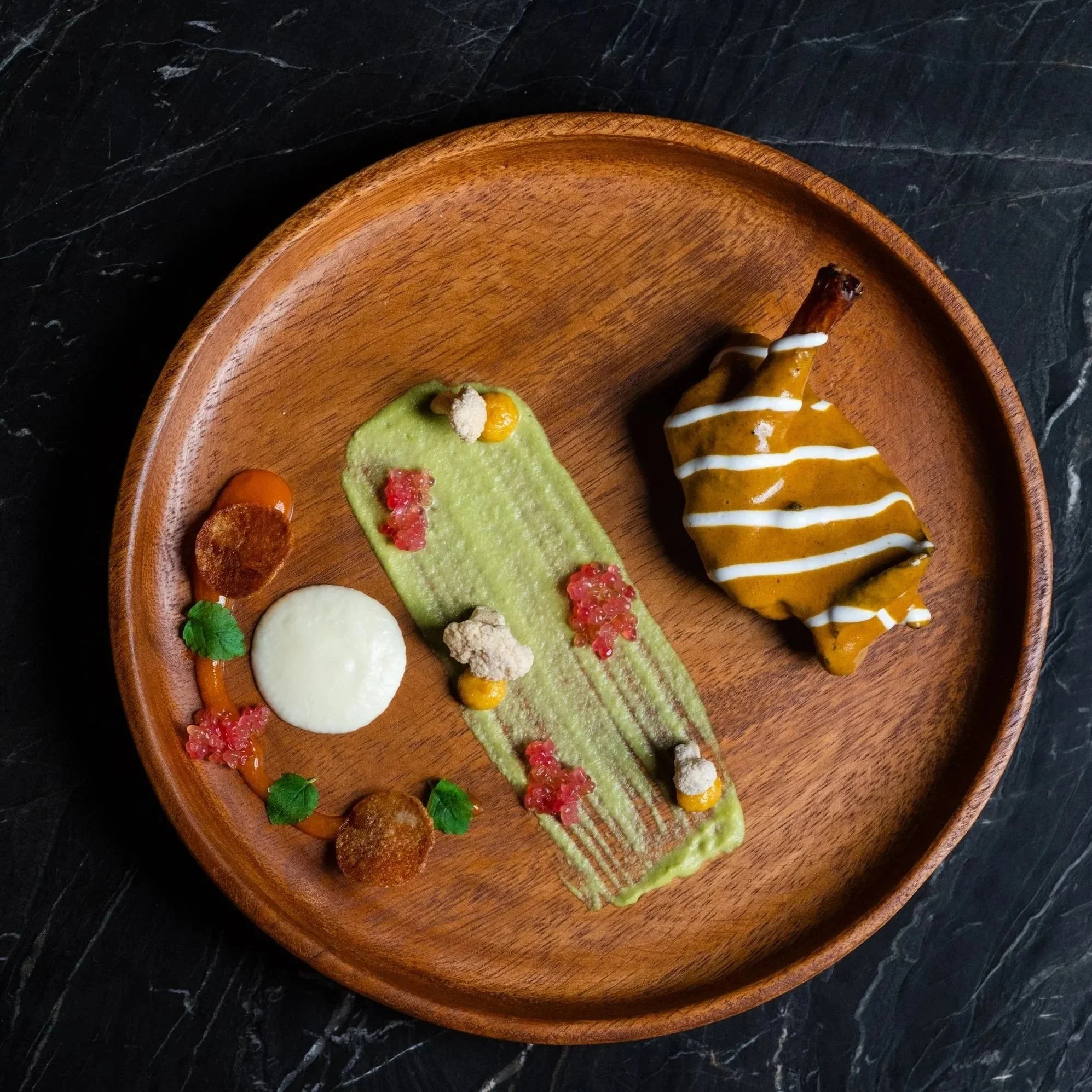 A tasting menu course plated on wood: green puree, pickled garnishes, and a glazed pastry.