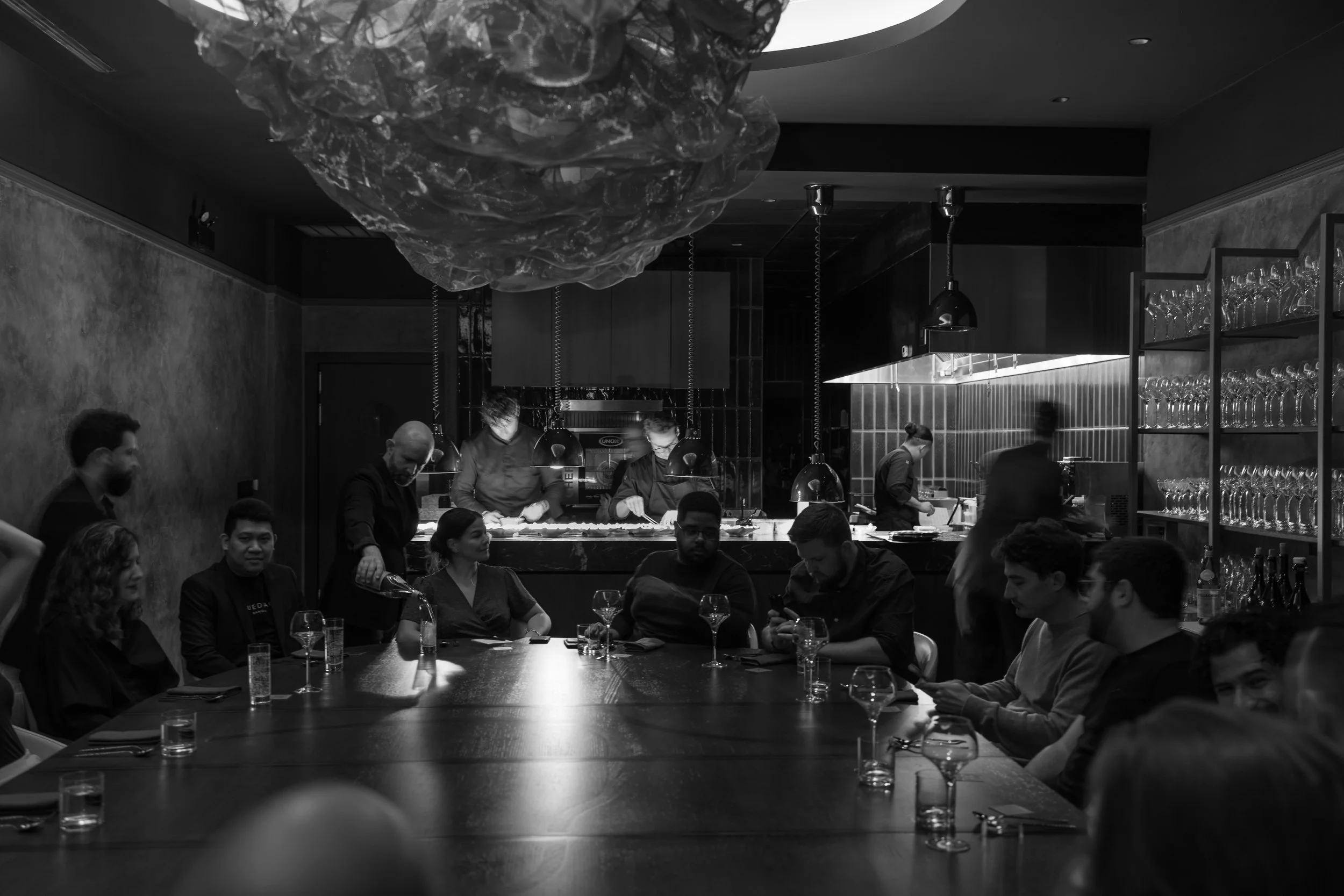 The Class Act chefs at work behind the communal table while guests dine, seen from the end of the room.