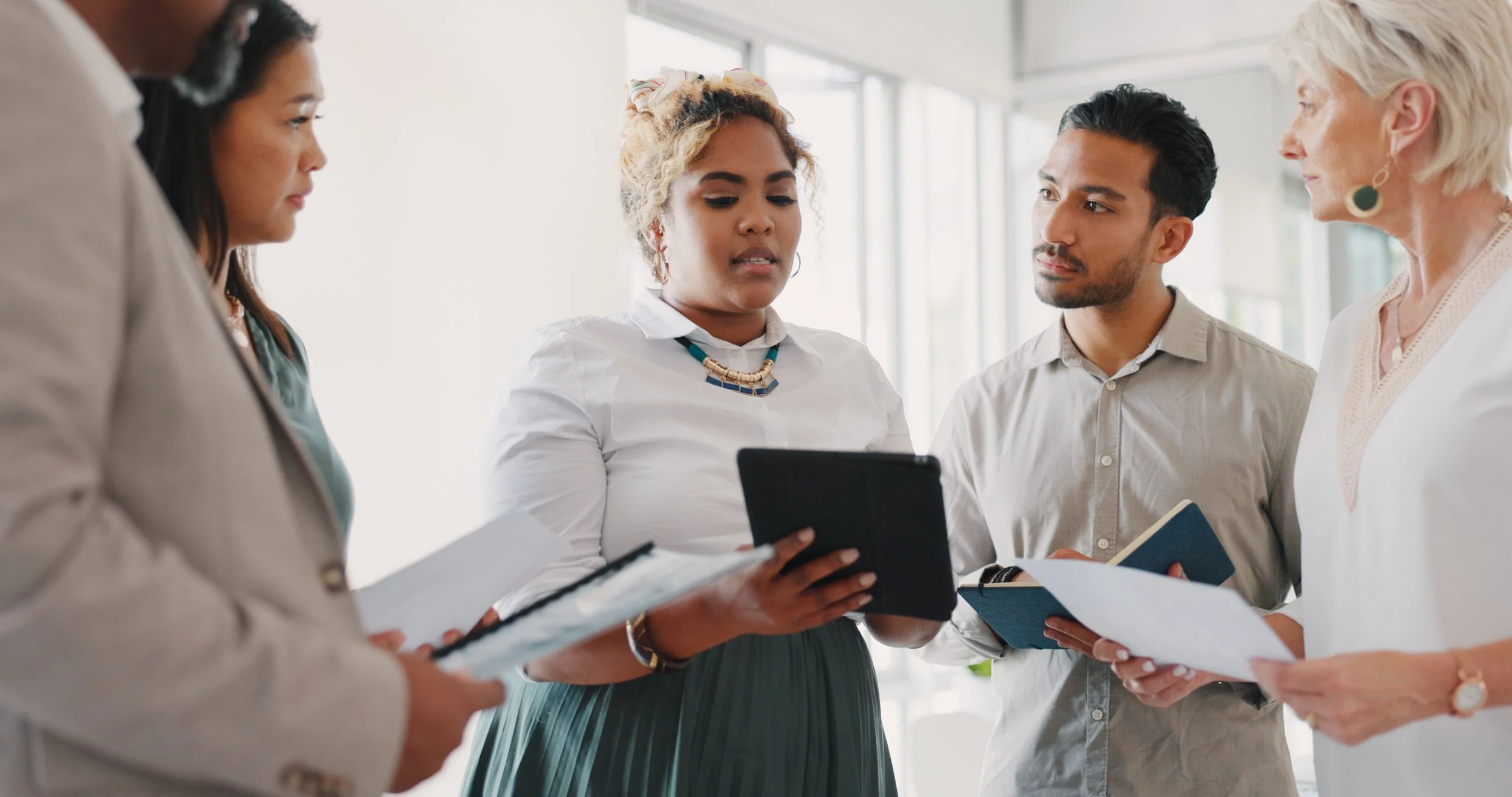 Five diverse adults engaged in discussion, holding papers and notebooks, in a well-lit office space.