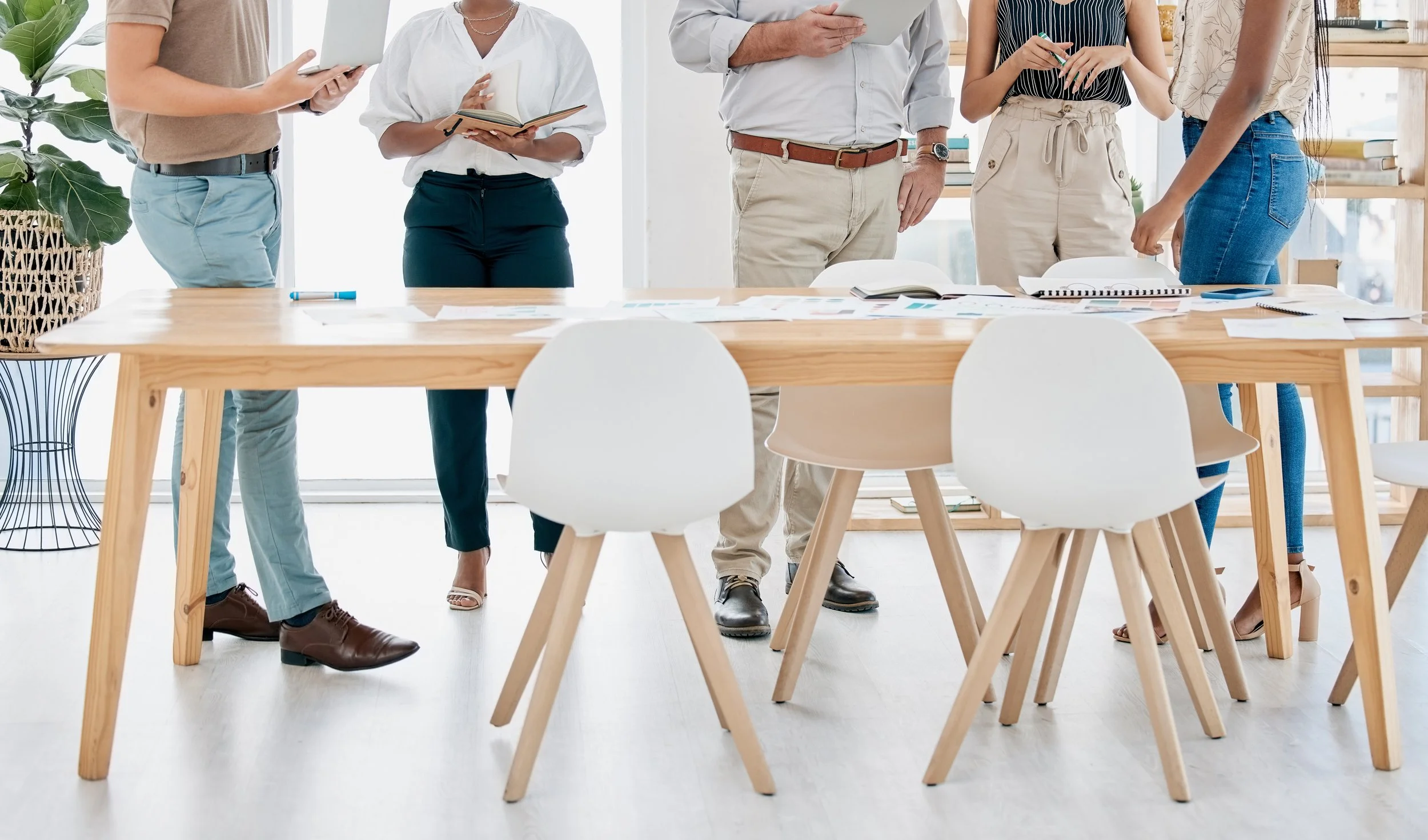 A group of five people standing around a wooden table in a bright office, engaged in discussion or collaboration, with documents and notebooks on the table.